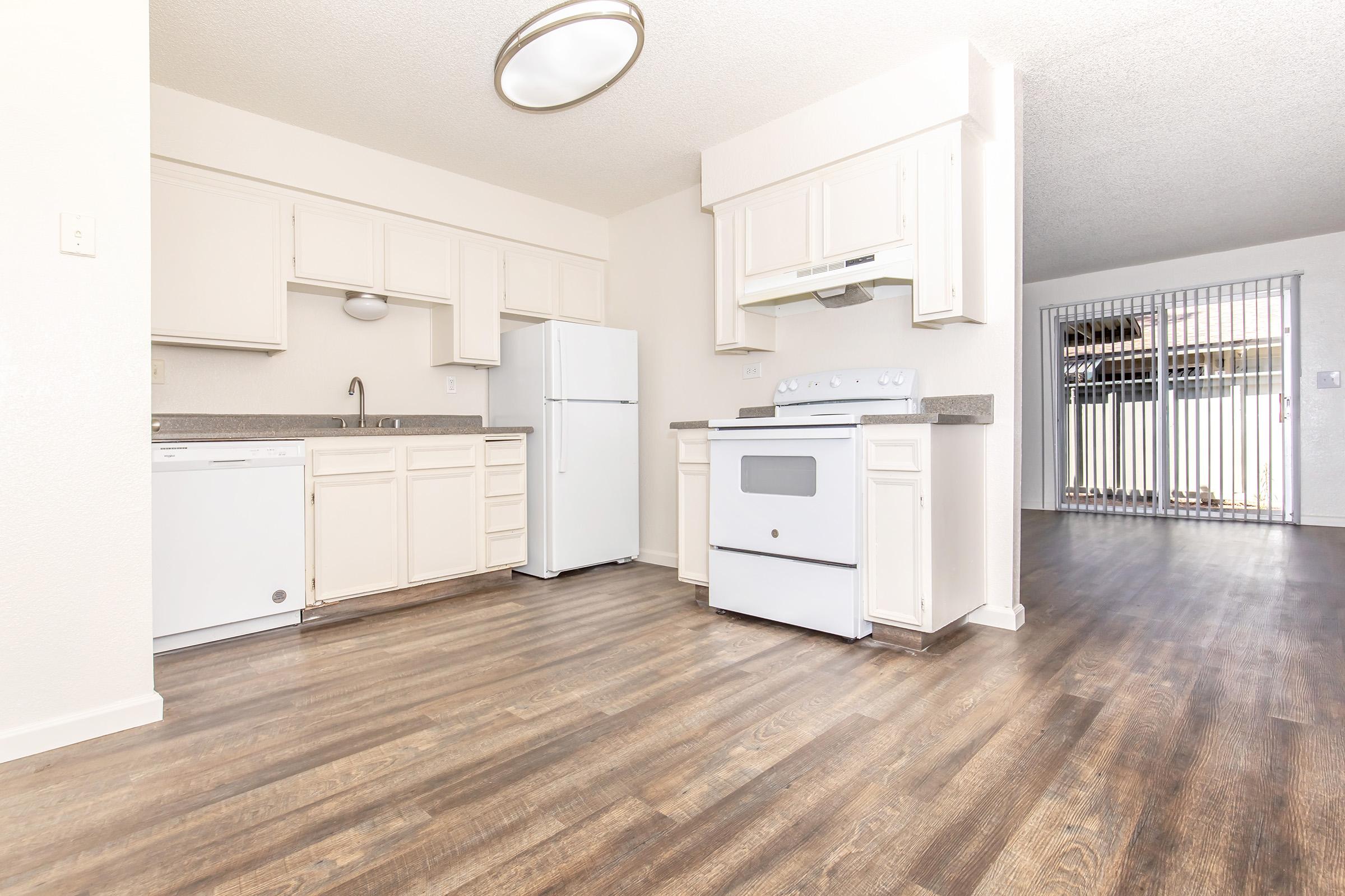 A modern kitchen featuring white cabinetry, a refrigerator, a stove with an oven, and a dishwasher. The space has a wood-like floor and is brightly lit. In the background, there is an open area with sliding glass doors leading to an outdoor space, partially visible behind a security gate.