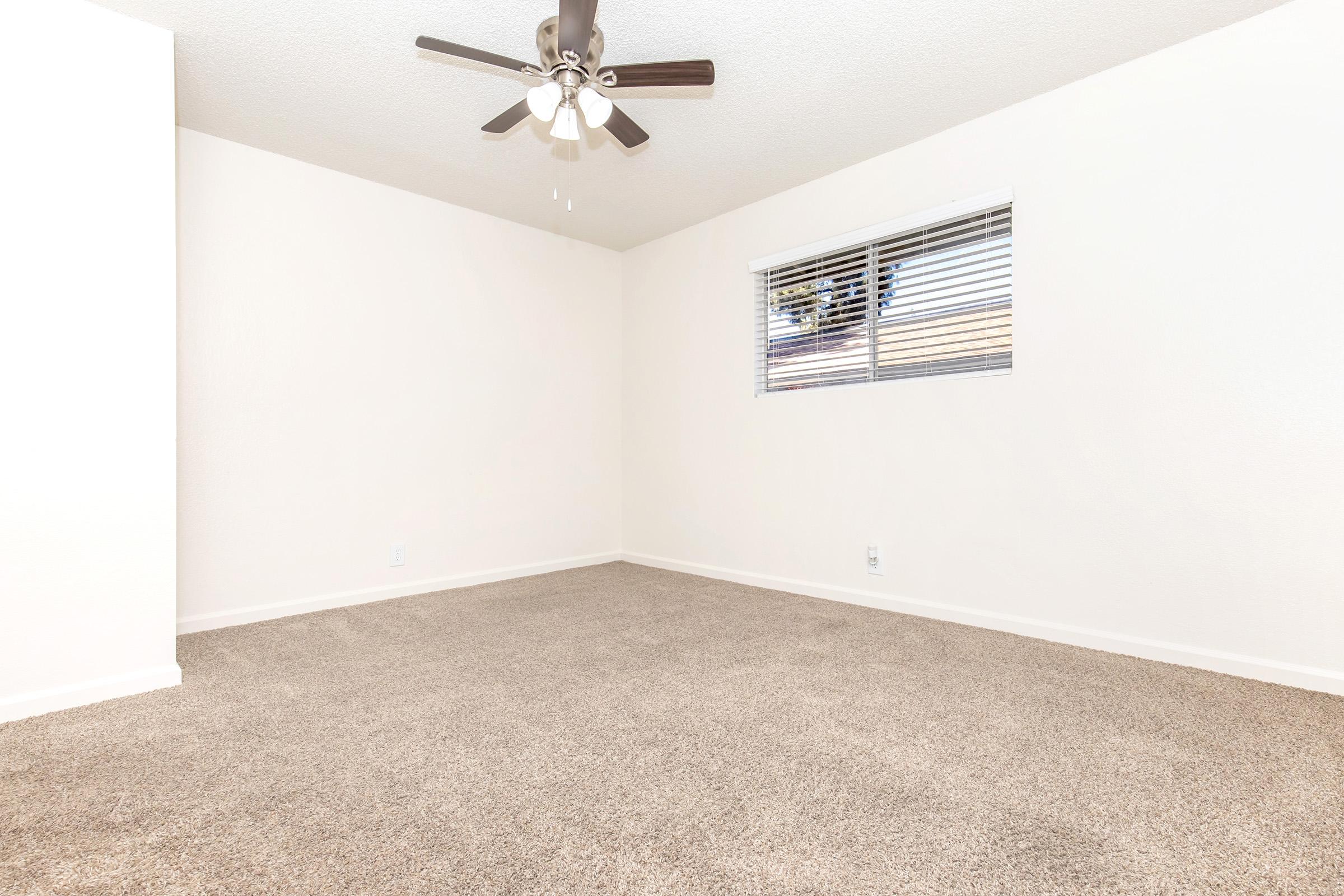 Empty room with light-colored walls and a window featuring blinds. The floor is covered with tan carpet, and a ceiling fan is visible above. The overall space appears bright and minimalistic, lacking furniture or decorations.