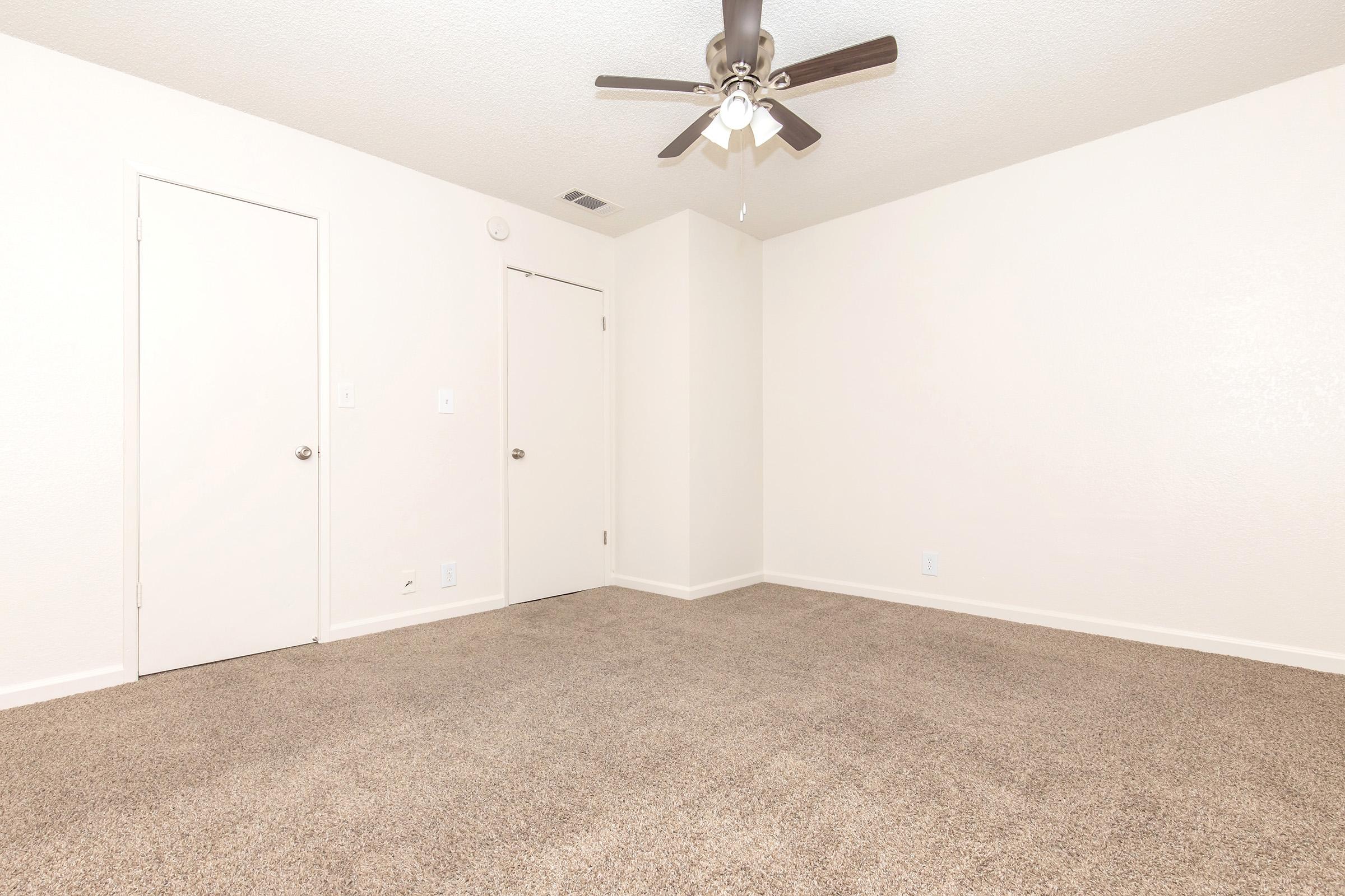 Empty room with light beige walls and carpeted flooring. A ceiling fan is mounted in the center, providing a modern touch. Two doors are visible on the left side, leading to an unknown space. The overall ambiance is bright and spacious, suitable for various personal or decorative styles.