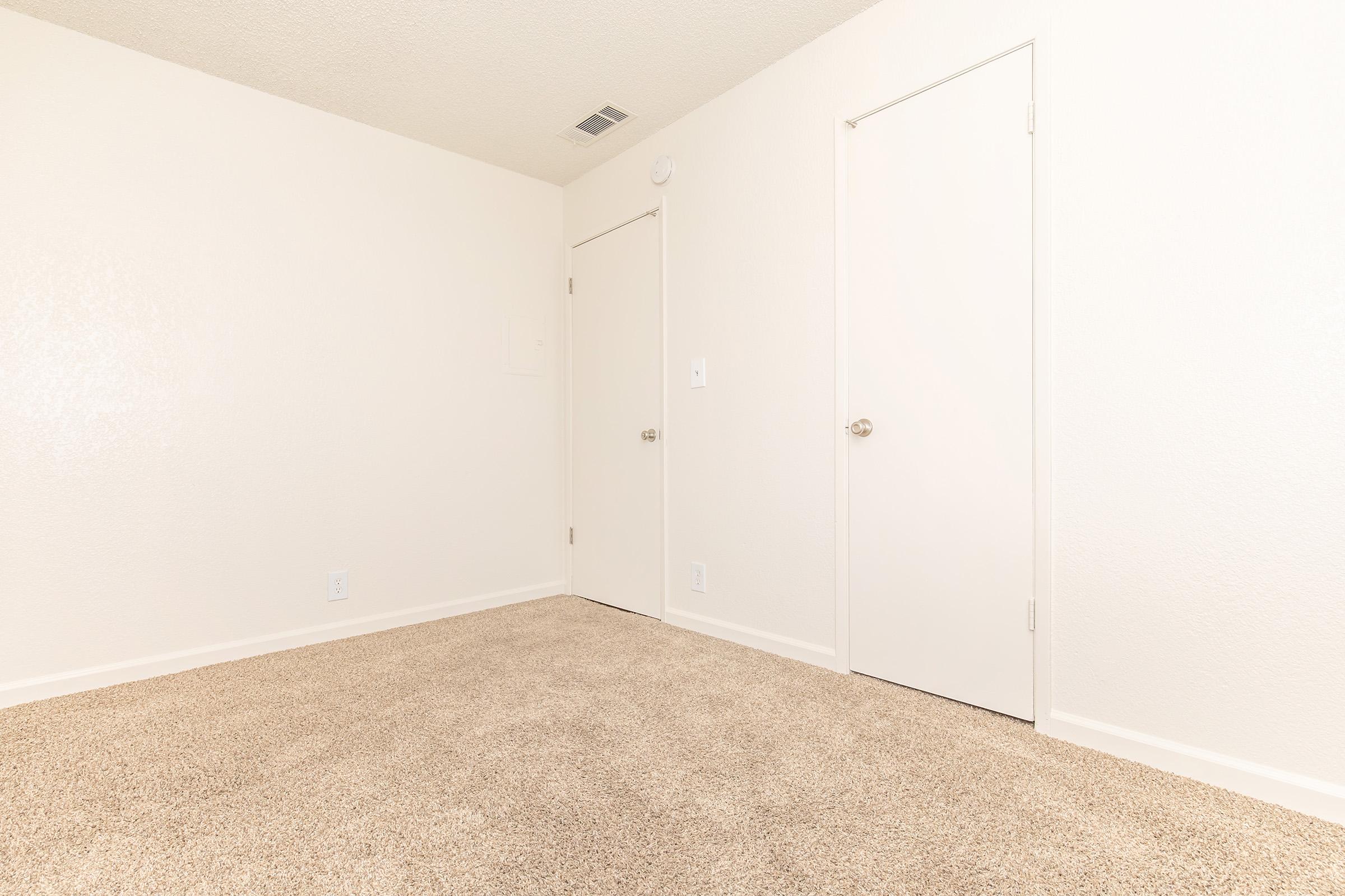 A clean, empty room featuring light-colored walls and beige carpet. There are two doors: one on the left and one on the right, both with silver handles. The space is well-lit and uncluttered, giving it a minimalist and airy feel.