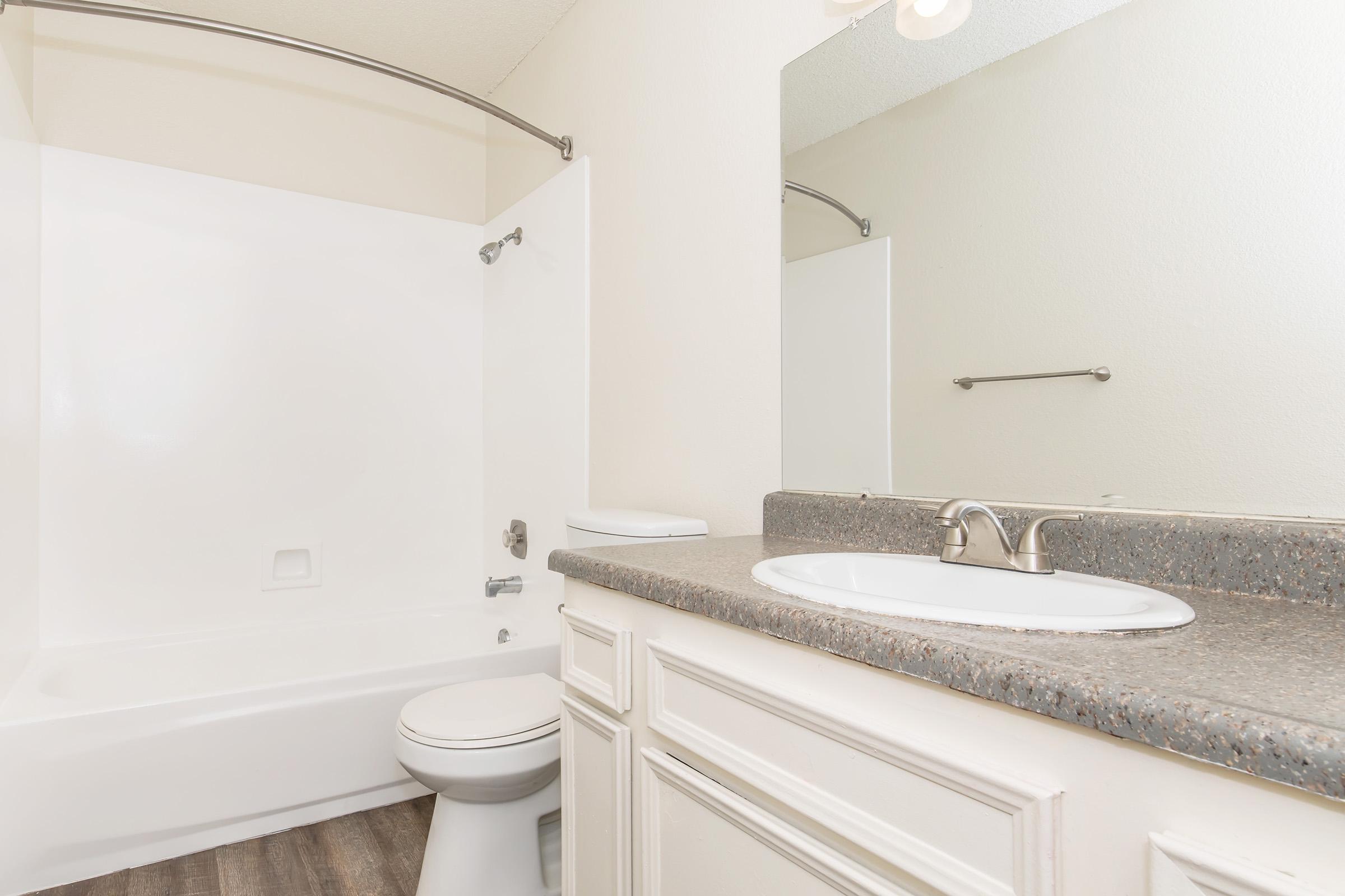 A clean and modern bathroom featuring a white bathtub and shower combination, a white toilet, and a countertop with a sink. The walls are light-colored, and the flooring appears to be a wood-like texture. A mirror hangs above the sink, and there is good lighting in the space.