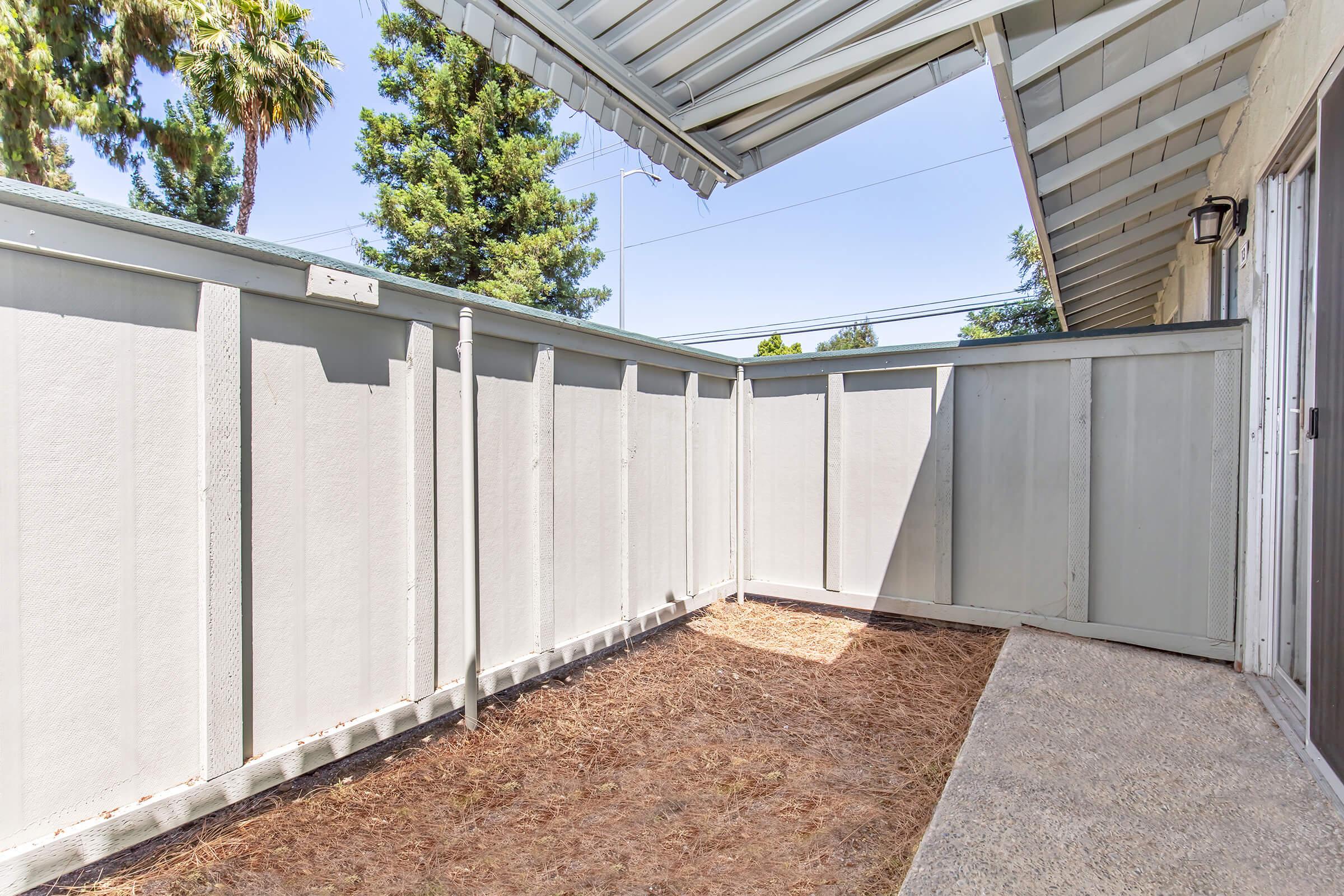 A small, enclosed outdoor space with gray wooden fencing, partially covered by a roof. The ground is bare and sandy, with a few patches of grass. Trees are visible in the background, and sunlight brightens the area, creating a serene atmosphere.