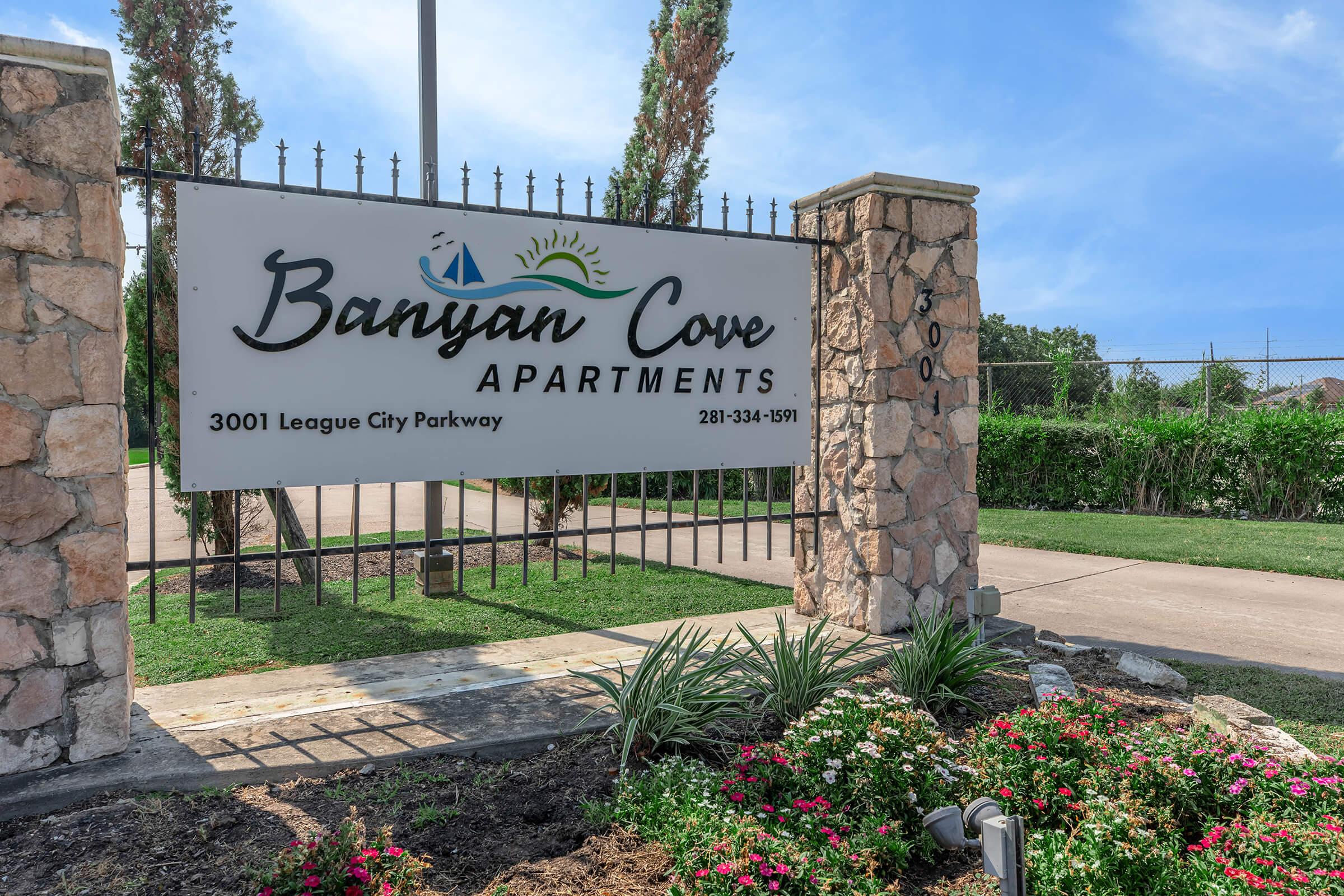 Sign for Banyan Cove Apartments located at 3001 League City Parkway, featuring a decorative stone structure, greenery, and colorful flowers in the foreground. The sign includes the apartment name and a contact number.
