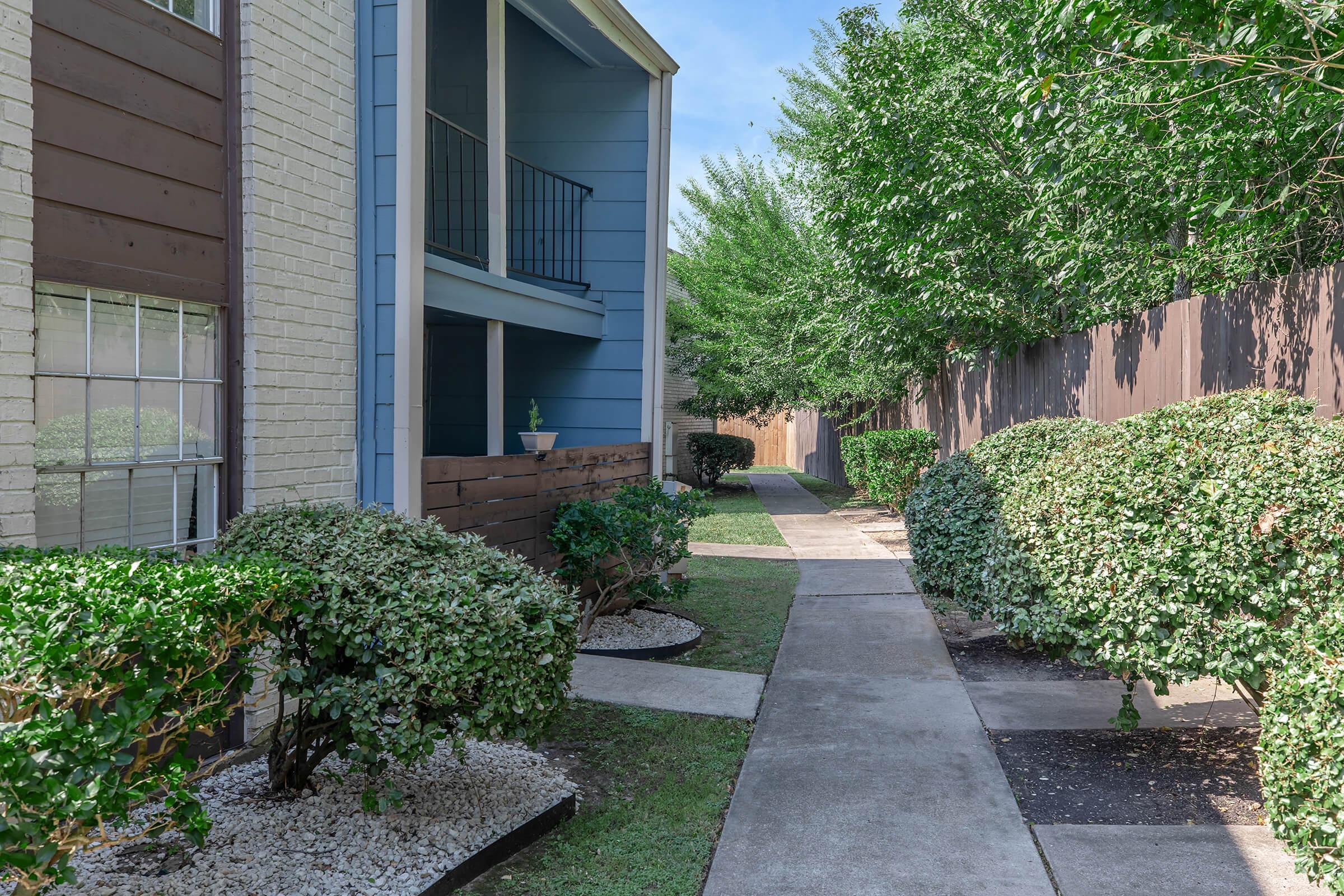 A pathway lined with well-maintained shrubs and small decorative rocks, leading between two residential buildings. One building features a blue balcony and the other has a light-colored brick facade. Lush green trees are visible in the background under a clear blue sky.