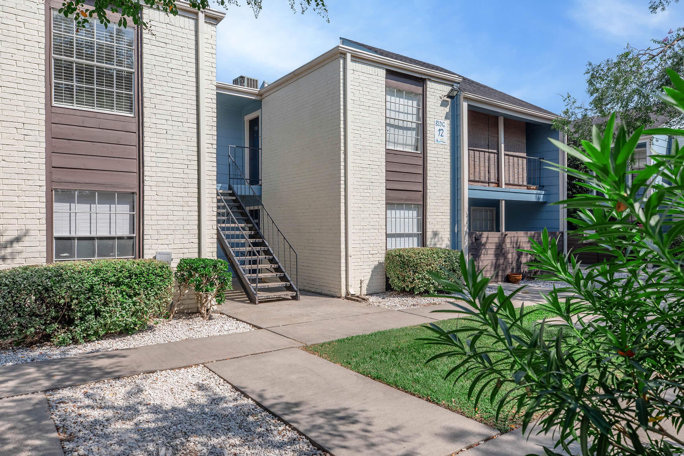 Image of an apartment building with two stories. The exterior features a mix of white and brown siding, a staircase leading to the second floor, and landscaped areas with shrubs and gravel. A pathway runs in front of the building, and clear blue skies are visible above.