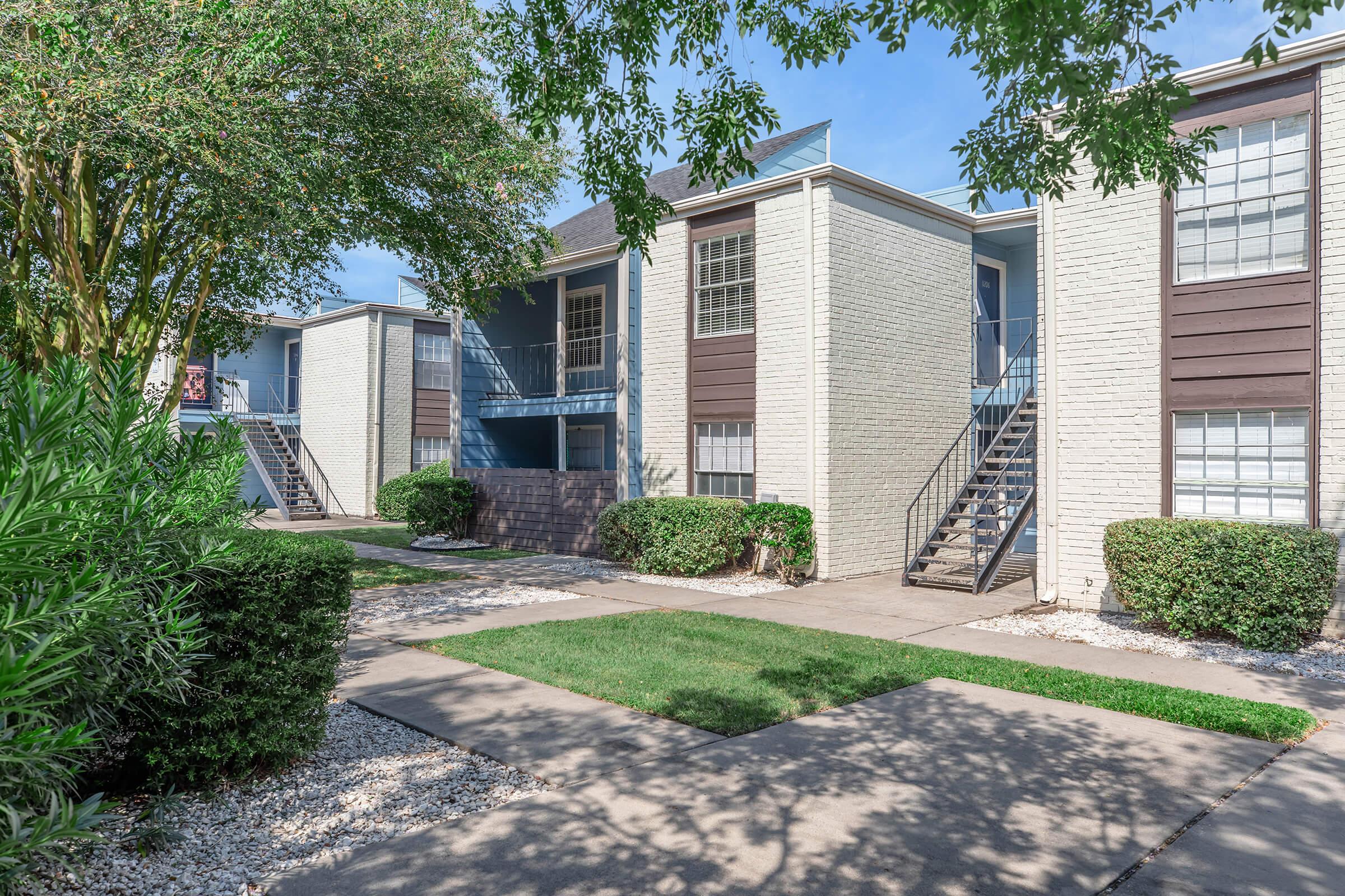 A view of a well-maintained multi-unit apartment complex featuring two-story buildings with brick and siding exteriors. Lush greenery surrounds the walkways and stairs, creating a welcoming atmosphere. The scene is set under a clear blue sky with soft sunlight illuminating the buildings.