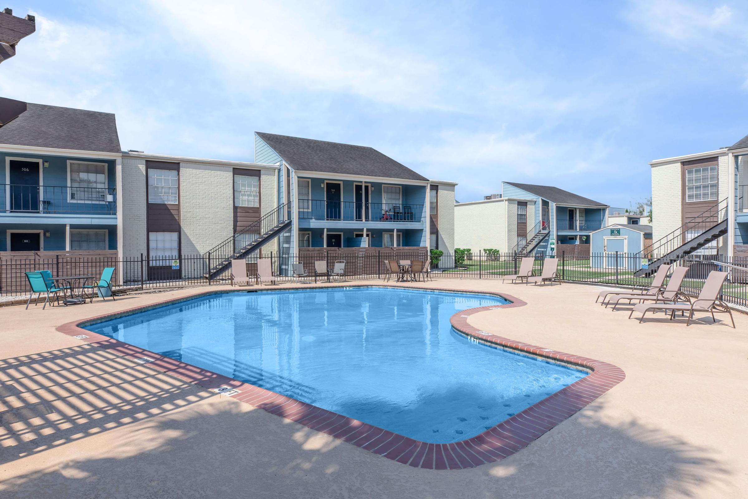 A clear outdoor swimming pool surrounded by lounge chairs, located in a residential complex. Two-story buildings with balconies can be seen in the background, under a bright blue sky. The area is well-maintained and inviting, perfect for relaxation and leisure activities.