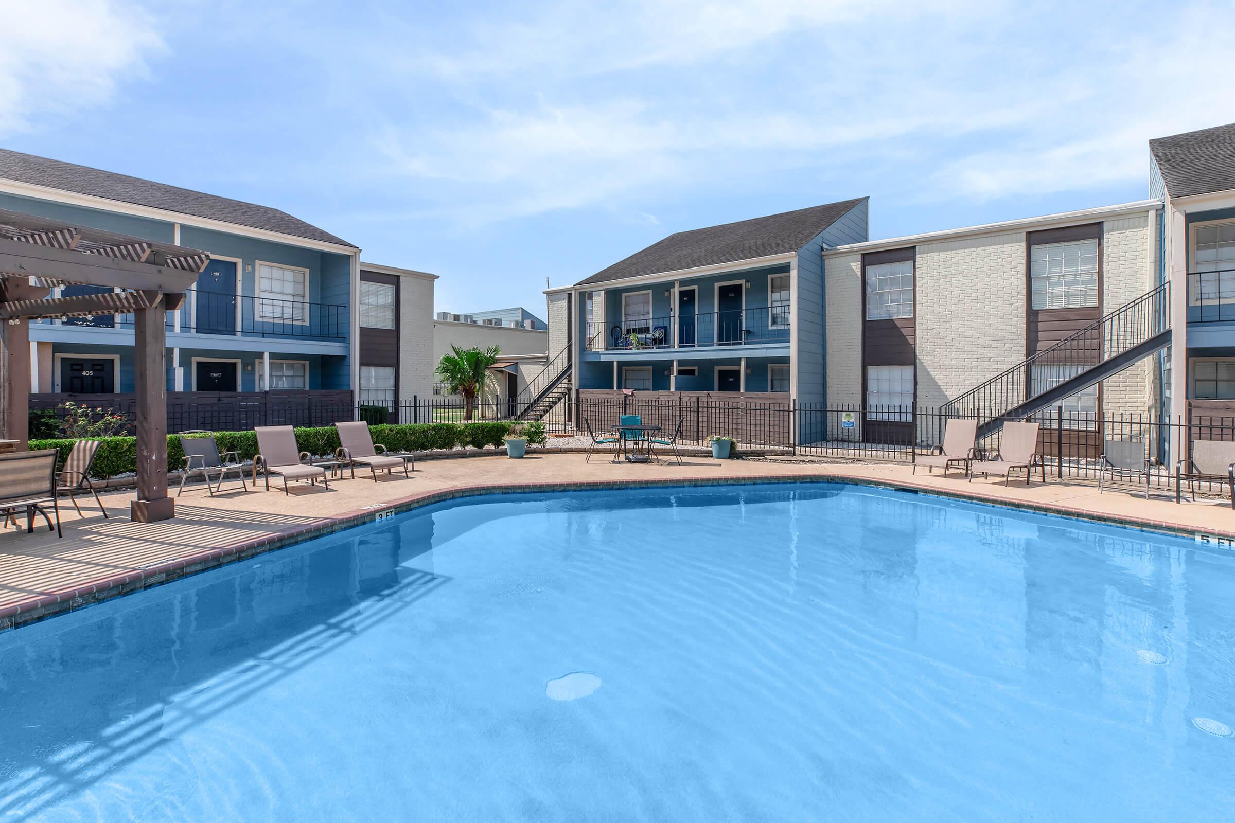 A clear swimming pool surrounded by lounge chairs and a gated area. In the background, there are two multi-story apartment buildings with balconies and a well-maintained landscape featuring palm trees. The sky is blue with a few clouds, giving a bright, inviting atmosphere.