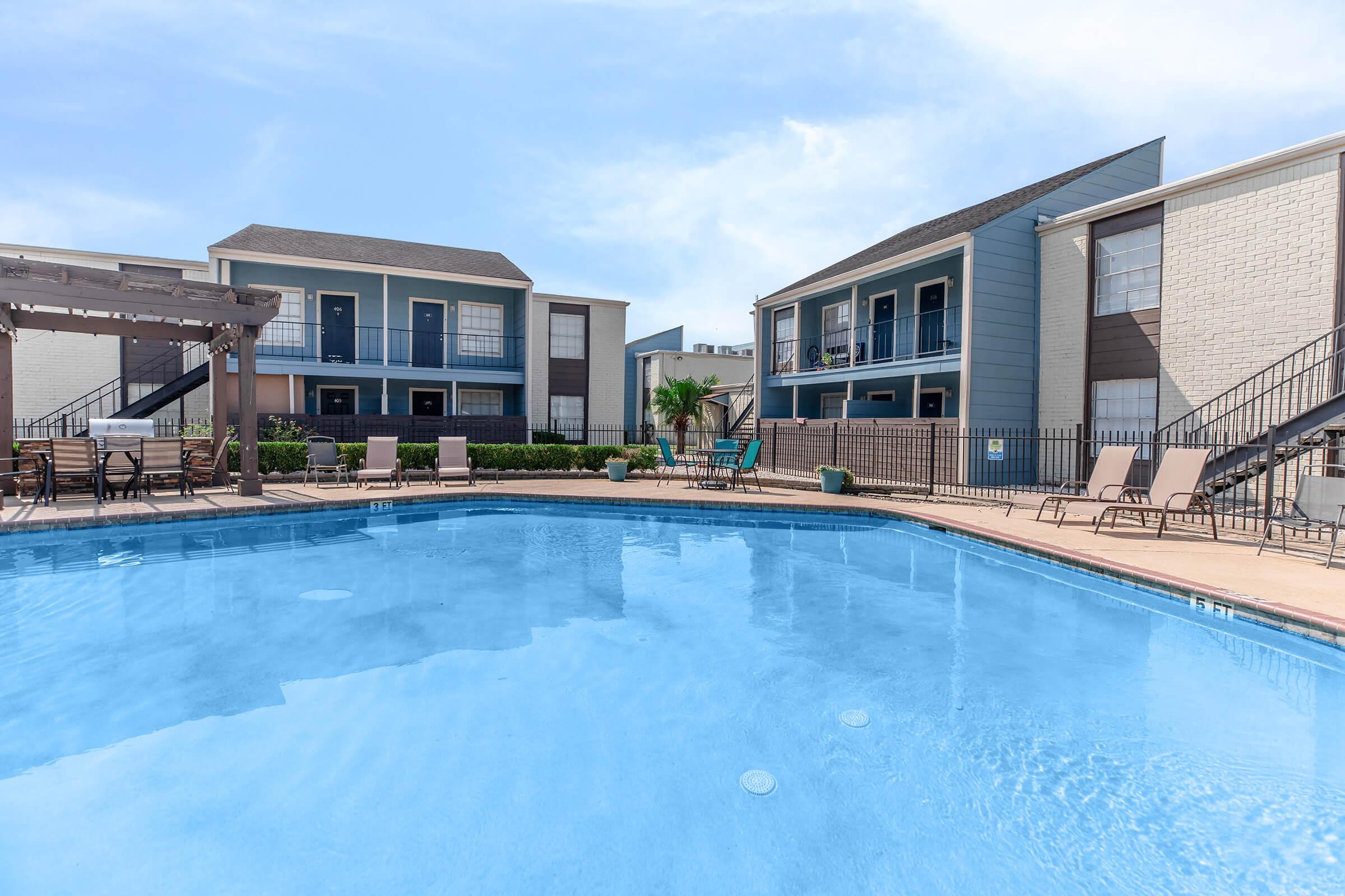 A clear blue swimming pool surrounded by lounge chairs, with two multi-story apartment buildings in the background. The area is enclosed by a fence, and there are some green plants and landscaping visible around the pool. The sky is bright and sunny.