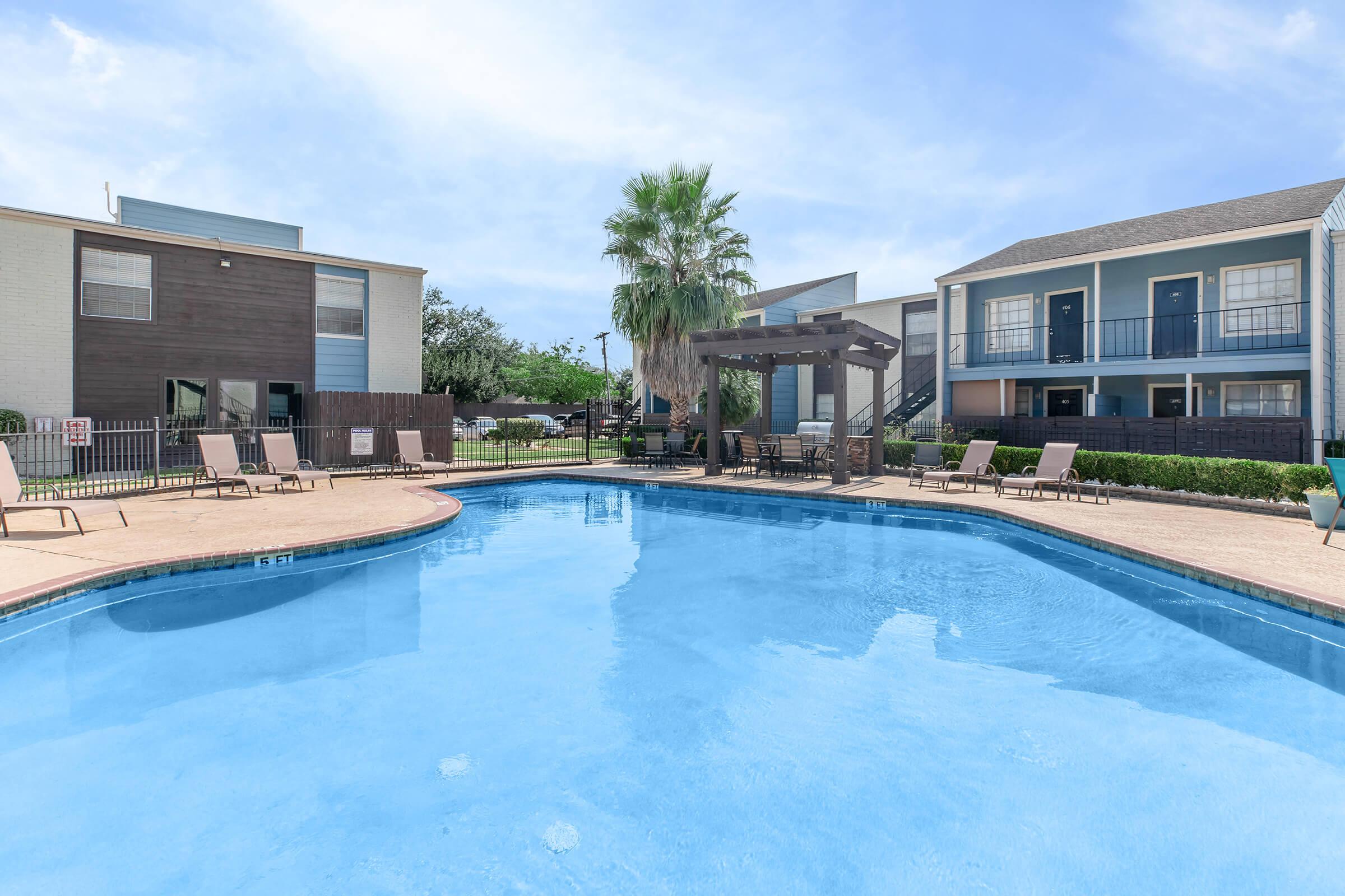 A bright, inviting swimming pool surrounded by lounge chairs and a shaded pergola. In the background, well-maintained apartment buildings and lush greenery create a relaxing atmosphere under a clear blue sky.