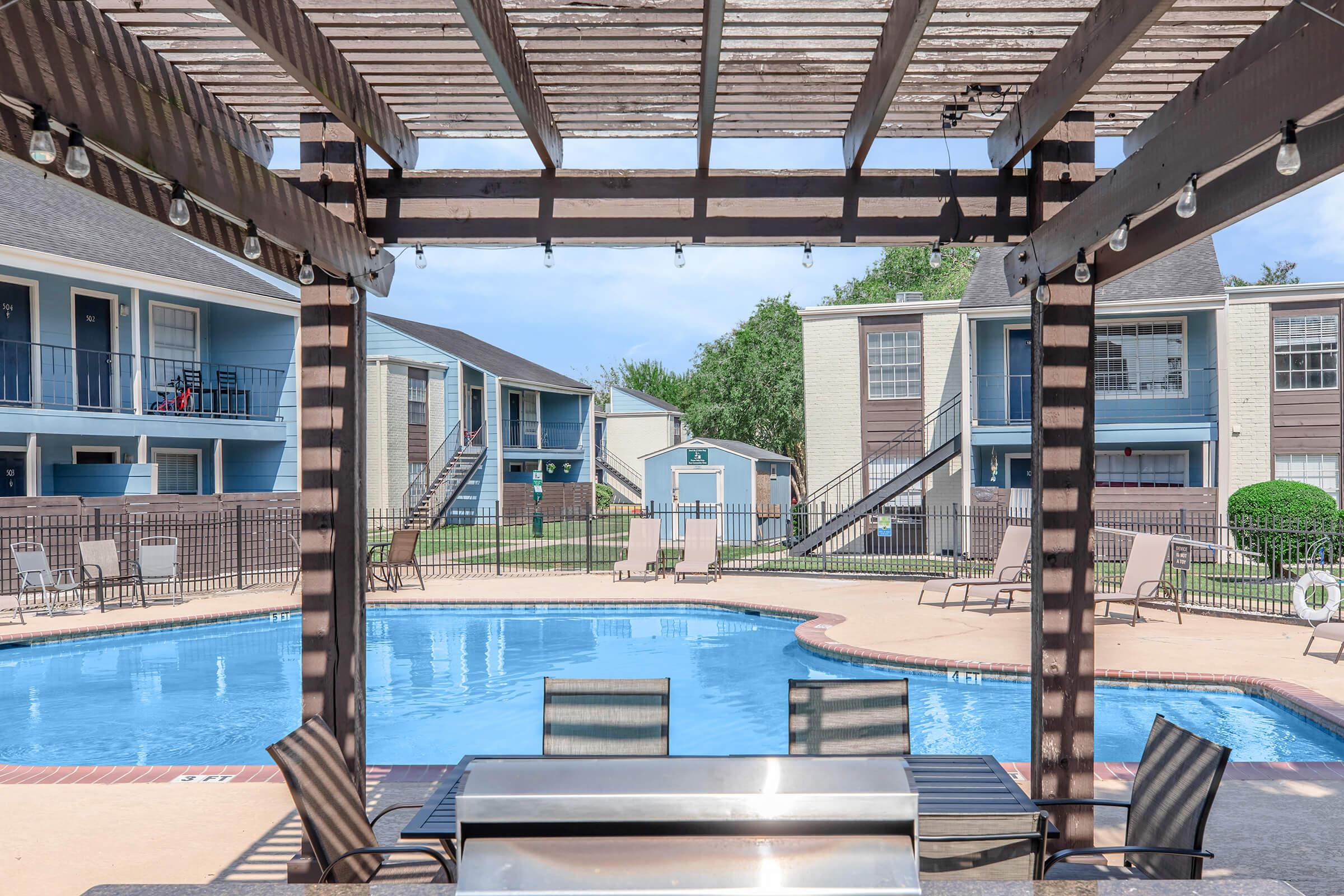 A swimming pool surrounded by a deck and lounge chairs, viewed from a shaded patio area with a grill. In the background, there are two apartment buildings and a storage shed, with landscaping and clear blue skies.
