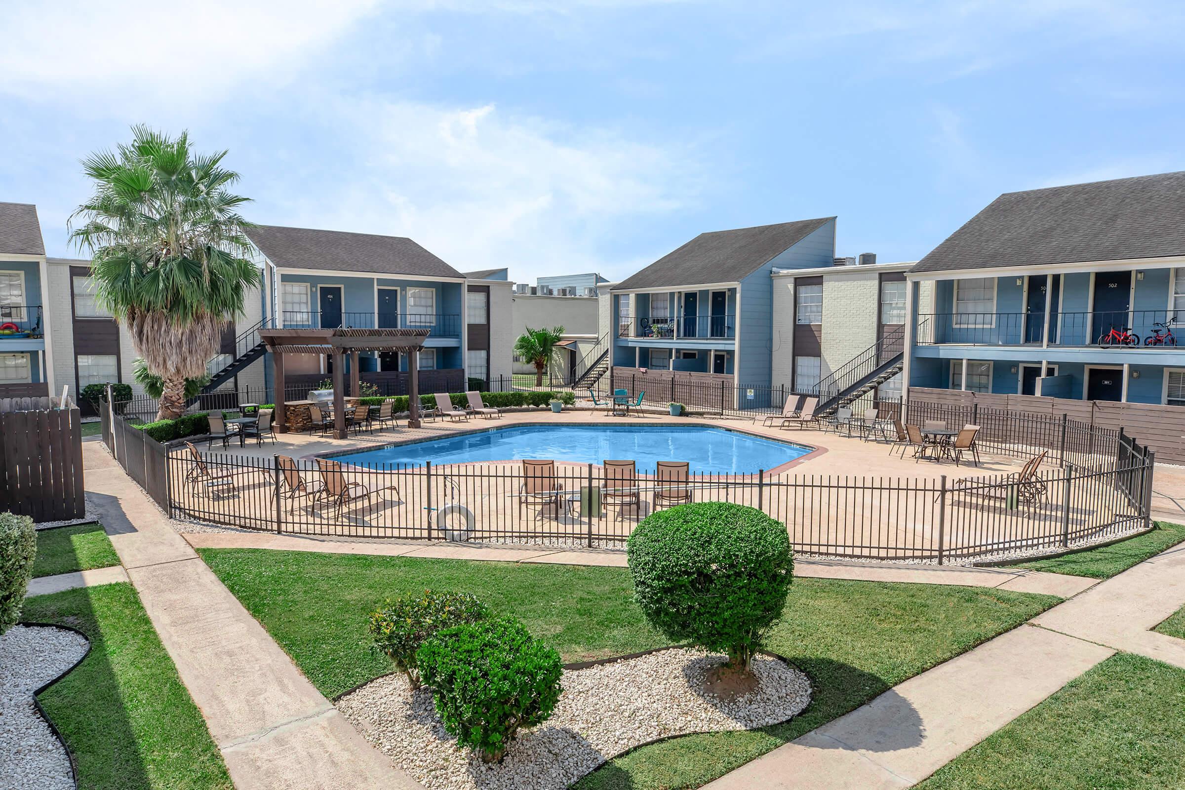 A view of an apartment complex featuring a central swimming pool surrounded by lounge chairs. The area is enclosed with a fence and landscaped with grass and shrubs. Several buildings are visible, with balconies overlooking the pool area. The sky is blue with a few clouds.