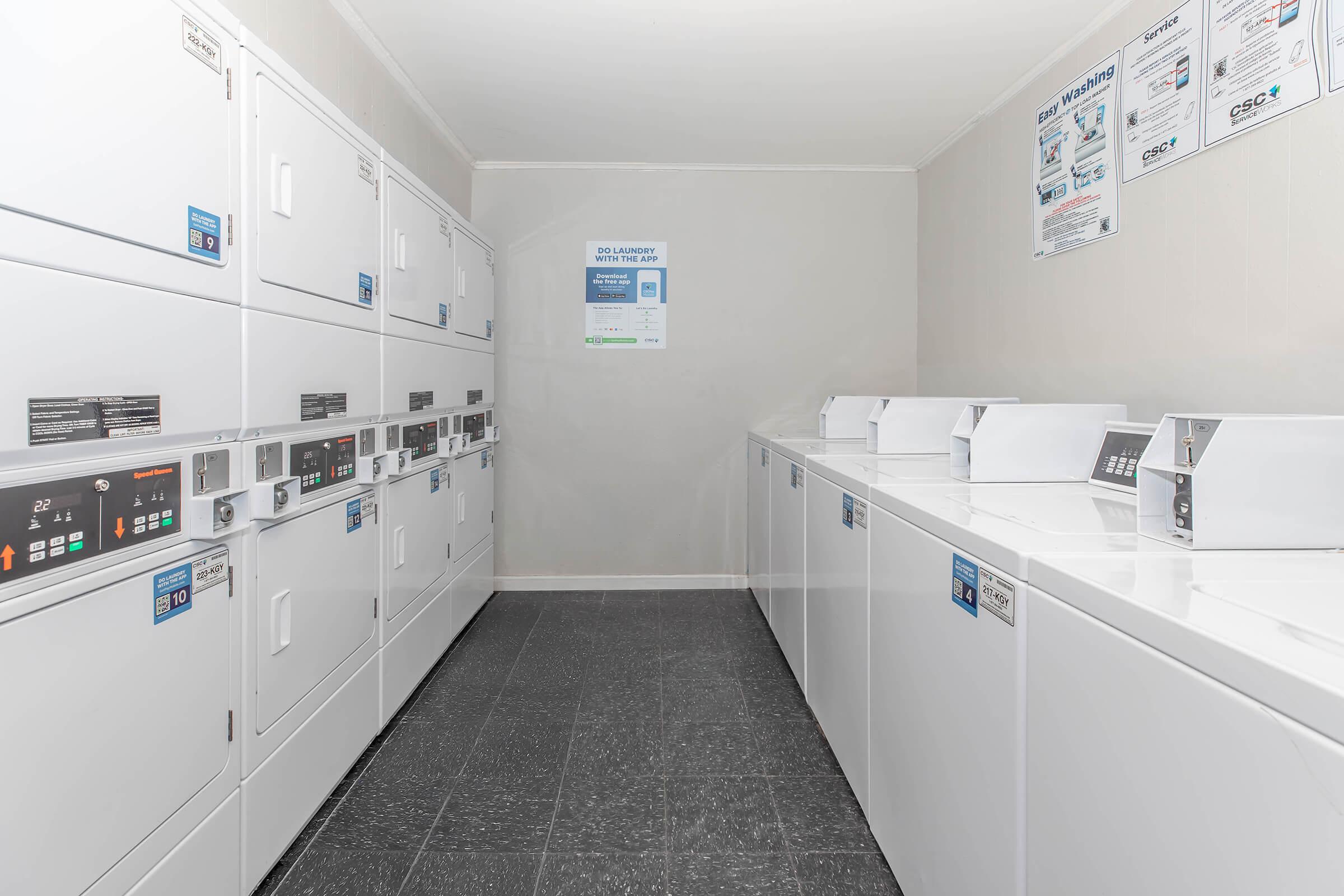A clean and modern laundry room featuring rows of white washing machines and dryers. The machines are equipped with control panels, and laundry instructions are displayed on the walls. The floor is dark and the walls are light-colored, creating a bright and spacious atmosphere.