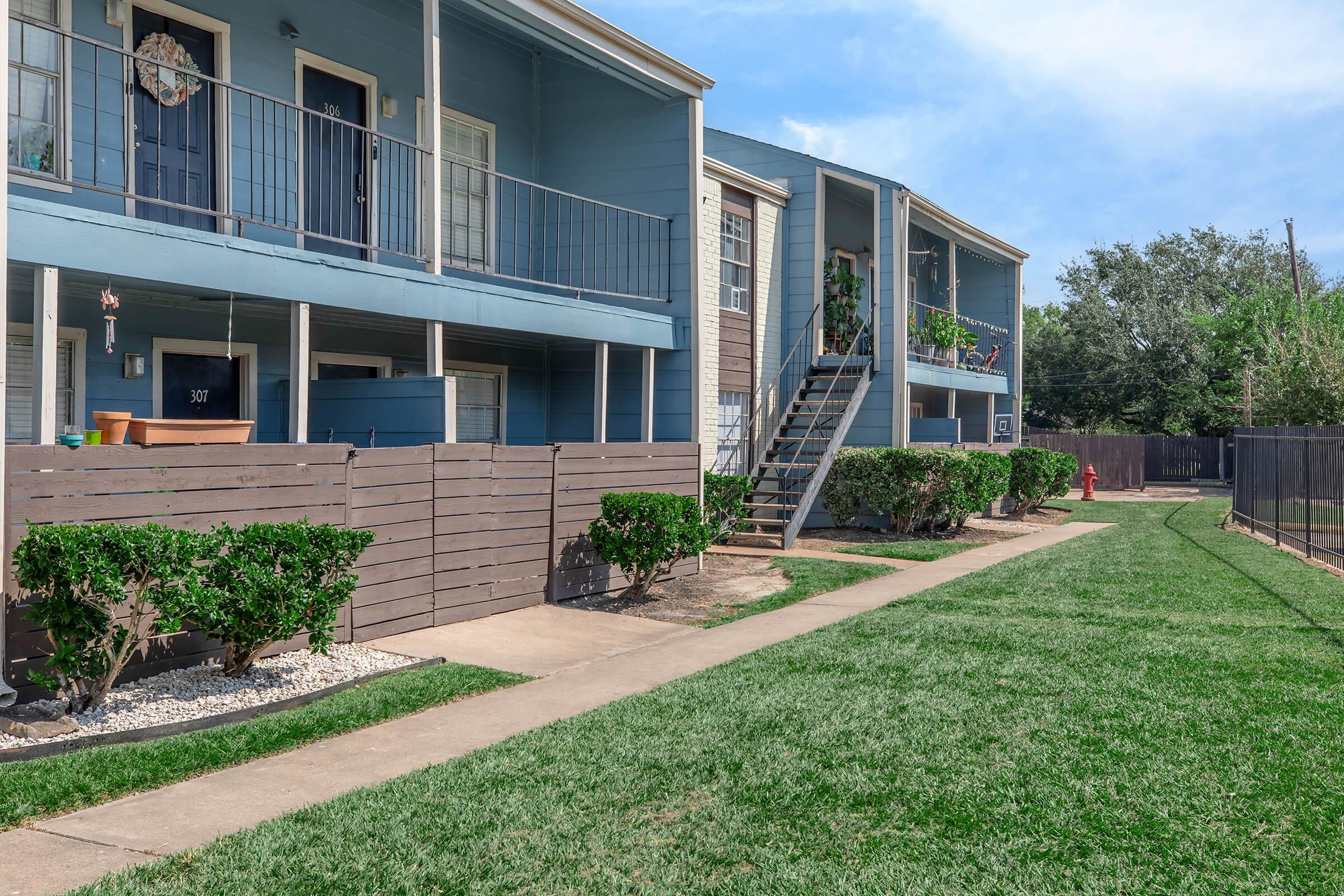 A view of a two-story apartment building with blue and brown siding. The walkway leads through a grassy area, bordered by low hedges. Each apartment has a small balcony with some plants. A staircase leads to the upper level on the right side. The scene is bright and well-maintained.