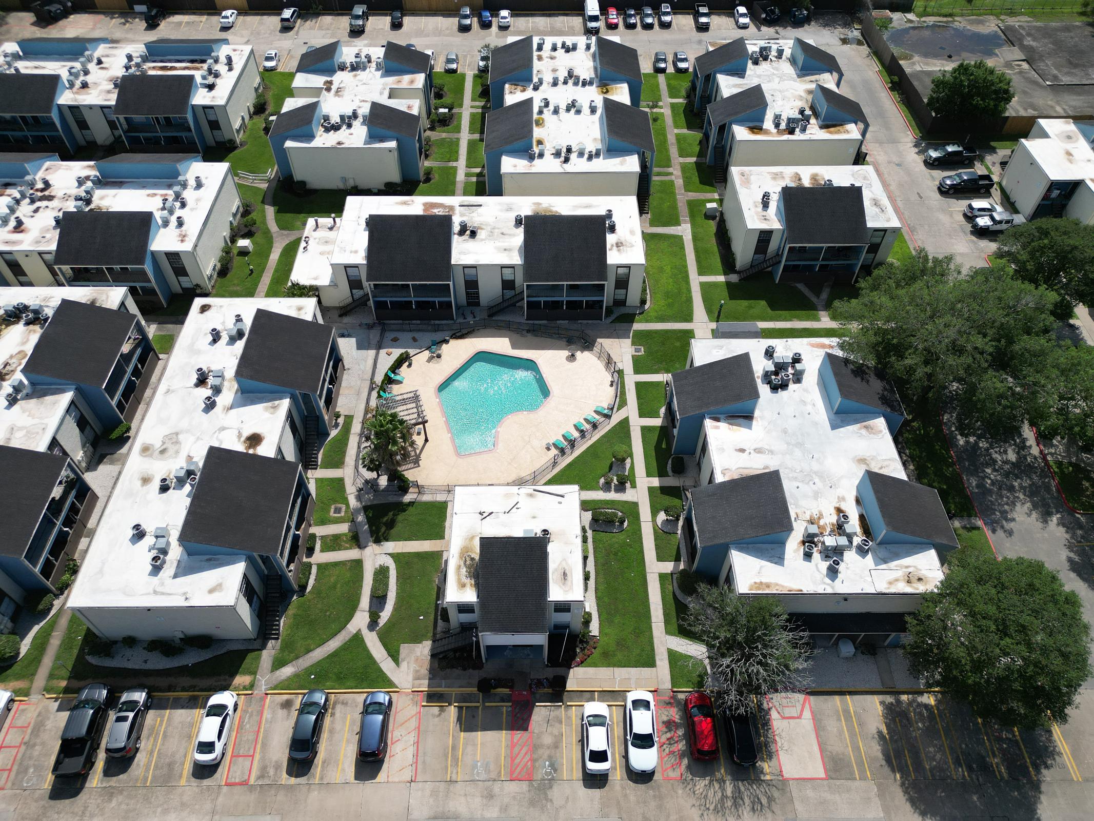 Aerial view of a residential complex featuring multiple two-story buildings arranged around a central swimming pool. The area includes landscaped green spaces and parking lots filled with vehicles. Several lounge chairs are positioned around the pool area.