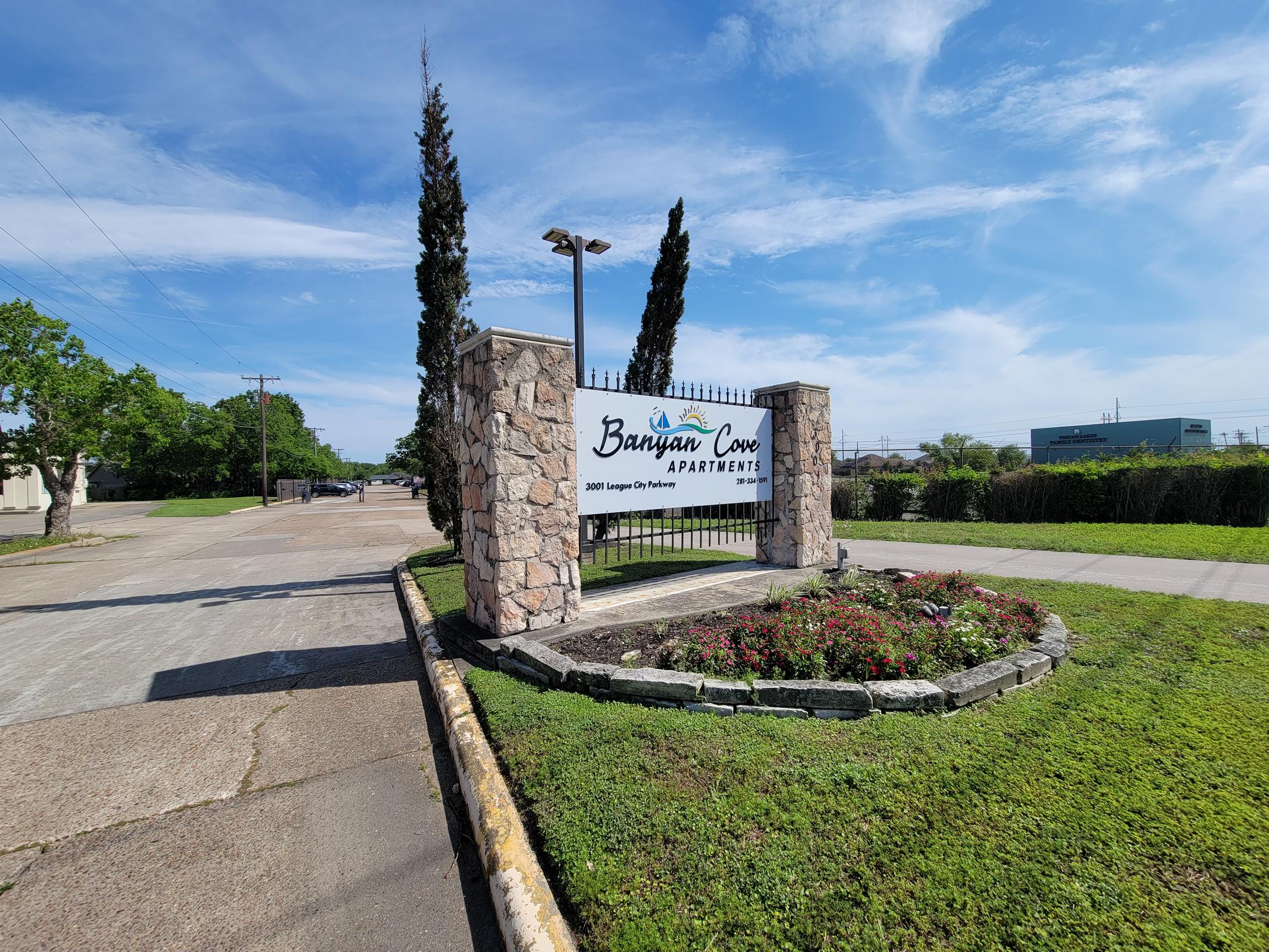 Sign for Banyan Cove Apartments with stone structure, landscaping featuring flowers, and a clear blue sky in the background. The sign includes the apartment name and address, surrounded by greenery and trees.