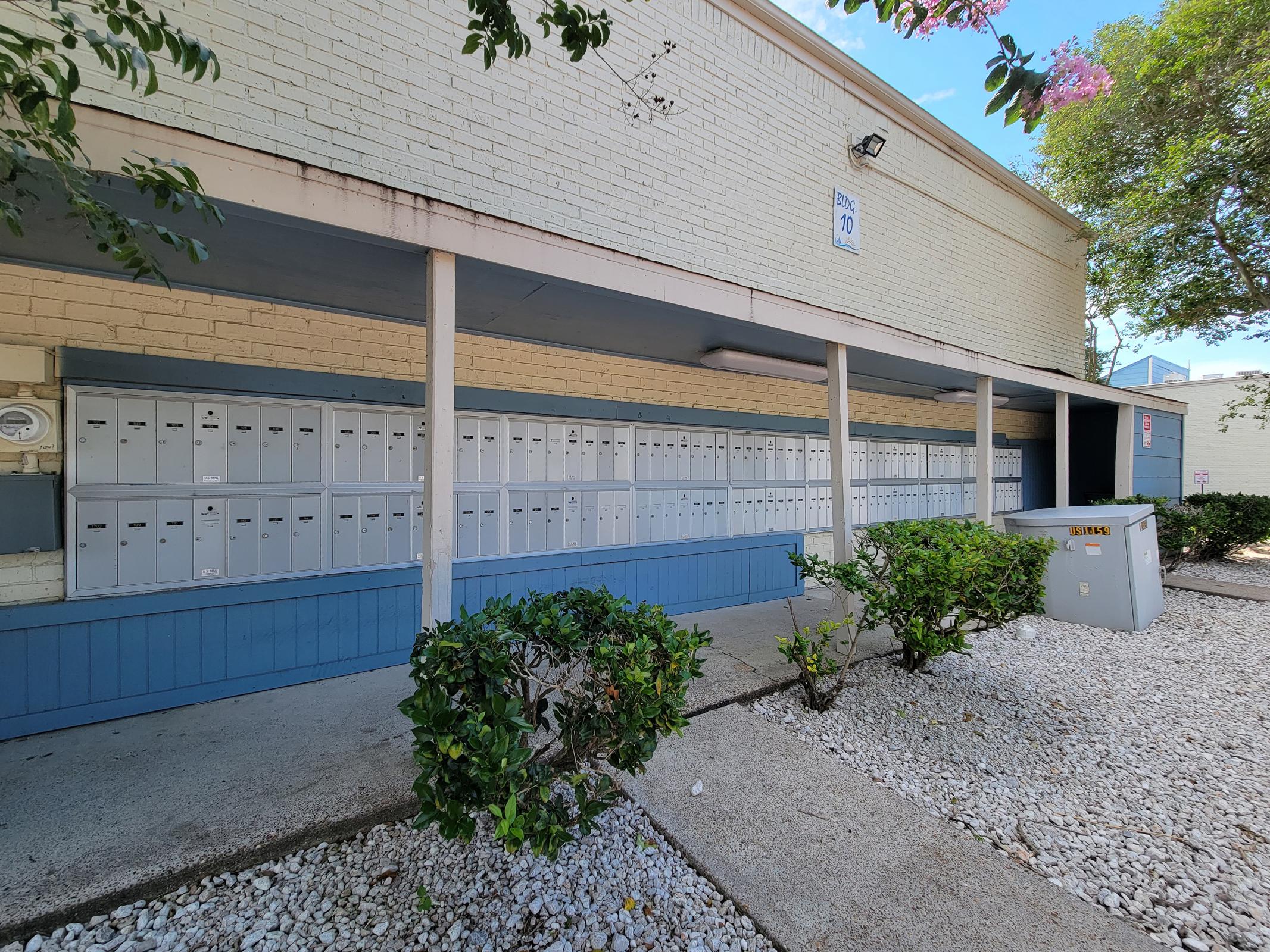 A row of gray mailboxes mounted on a wall, with a small utility box nearby. The area is surrounded by low shrubs and plants, and there is a clear blue sky above. The scene is well-lit and features a clean, tidy environment.