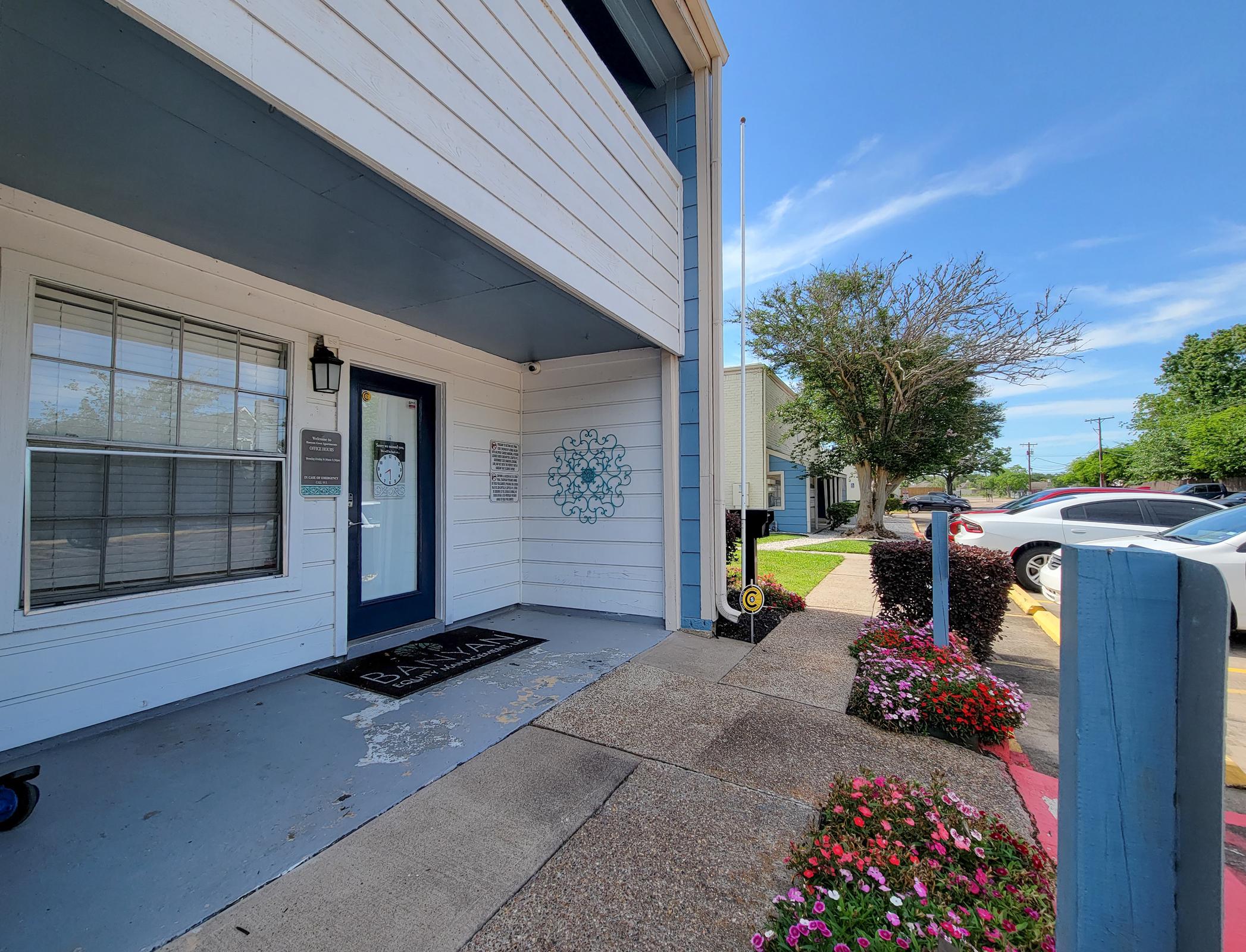 Exterior view of a building featuring a white wooden façade, a blue door, and a decorative wall piece. The entrance area includes a welcome mat and potted flowers. A parking lot is visible in the background with parked cars, and the sky is bright and clear.
