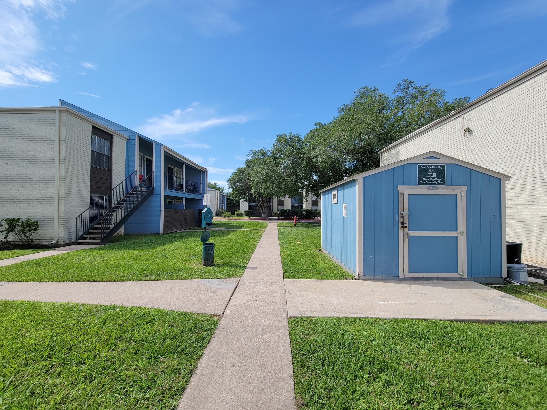 View of a residential area featuring two apartment buildings on either side of a walkway. The left building has stairs leading to upper floors. A blue shed is visible along the path, surrounded by green grass and trees under a clear blue sky.