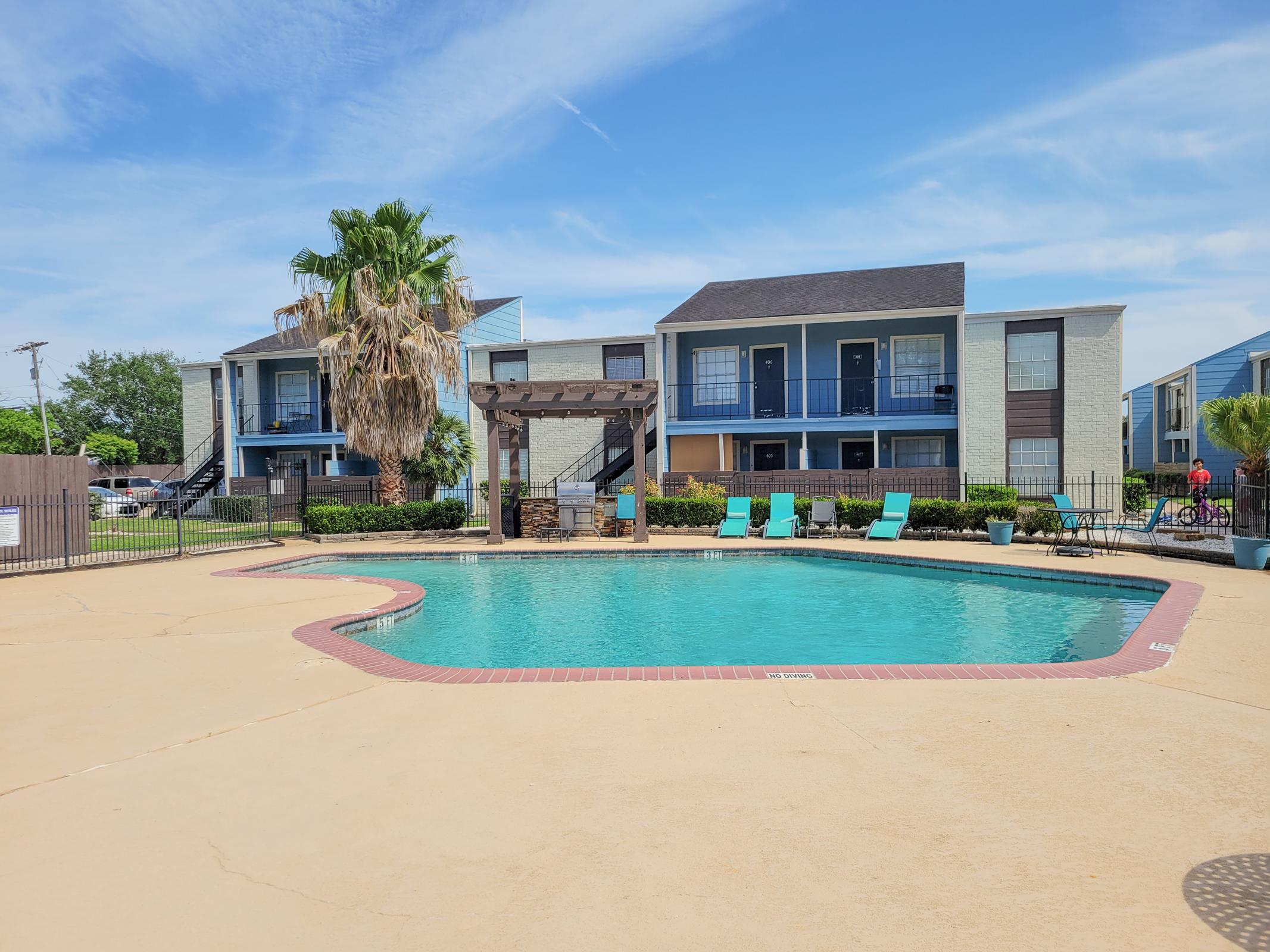 A serene apartment complex pool area featuring a clear blue pool surrounded by lounge chairs, palm trees, and manicured landscaping. Two multi-story buildings with balconies are visible in the background under a sunny sky.