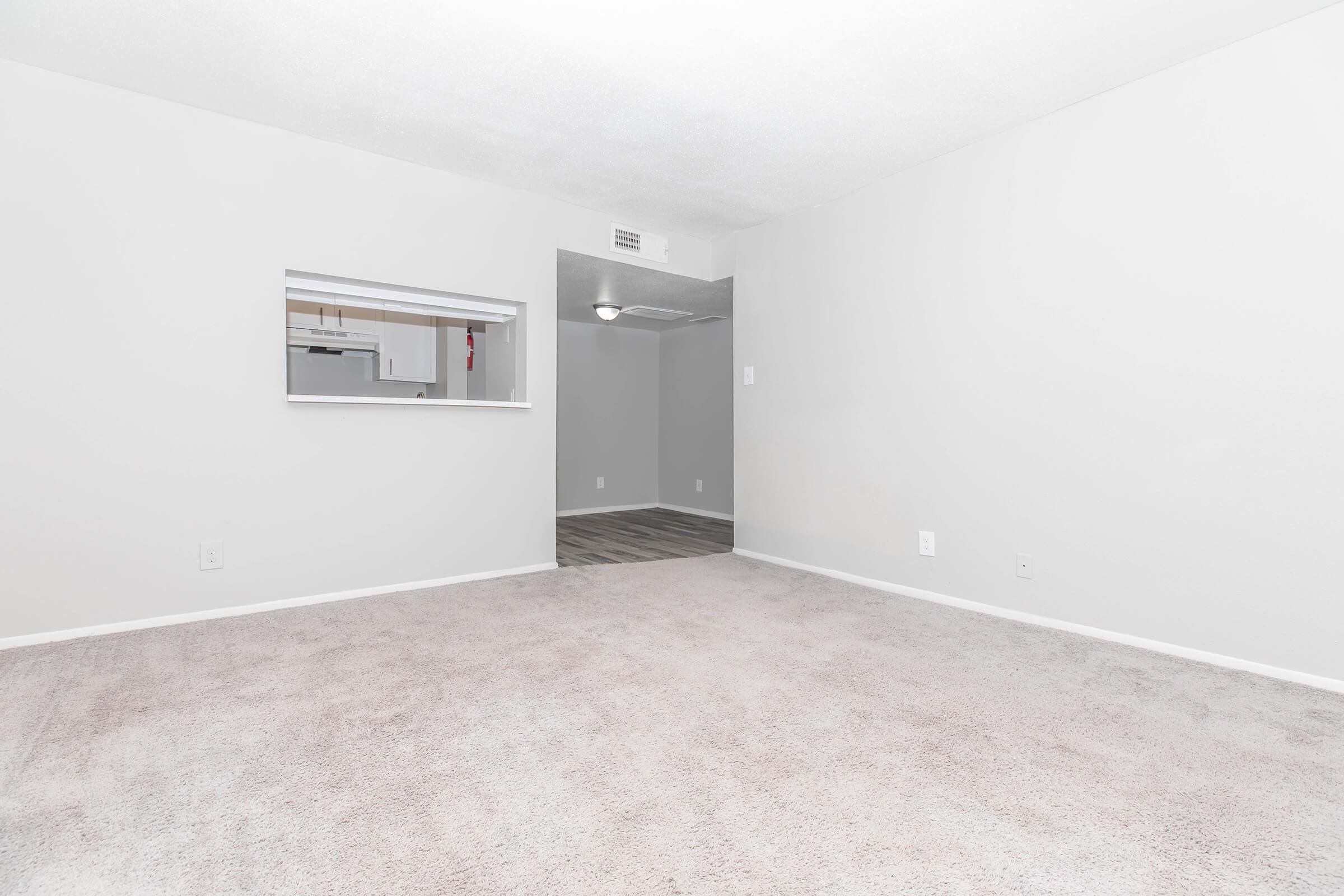 Spacious, empty living room with light gray walls and soft beige carpet. A large opening leads to a kitchen area visible in the background, featuring a light fixture. The overall atmosphere is bright and clean, inviting a sense of openness.