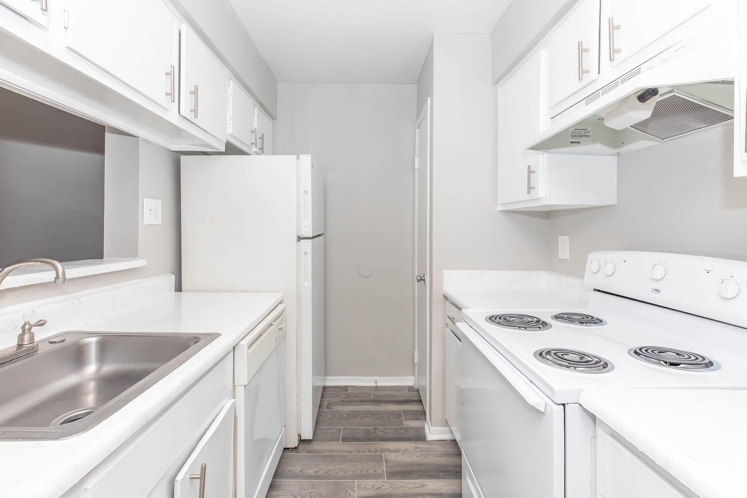 A bright, modern kitchen featuring white cabinetry, a stainless steel sink, and a white stove with four burners. A refrigerator is visible, along with a dishwasher next to the sink. The walls are painted a light gray, and the flooring is dark wood laminate, creating a clean and contemporary look.