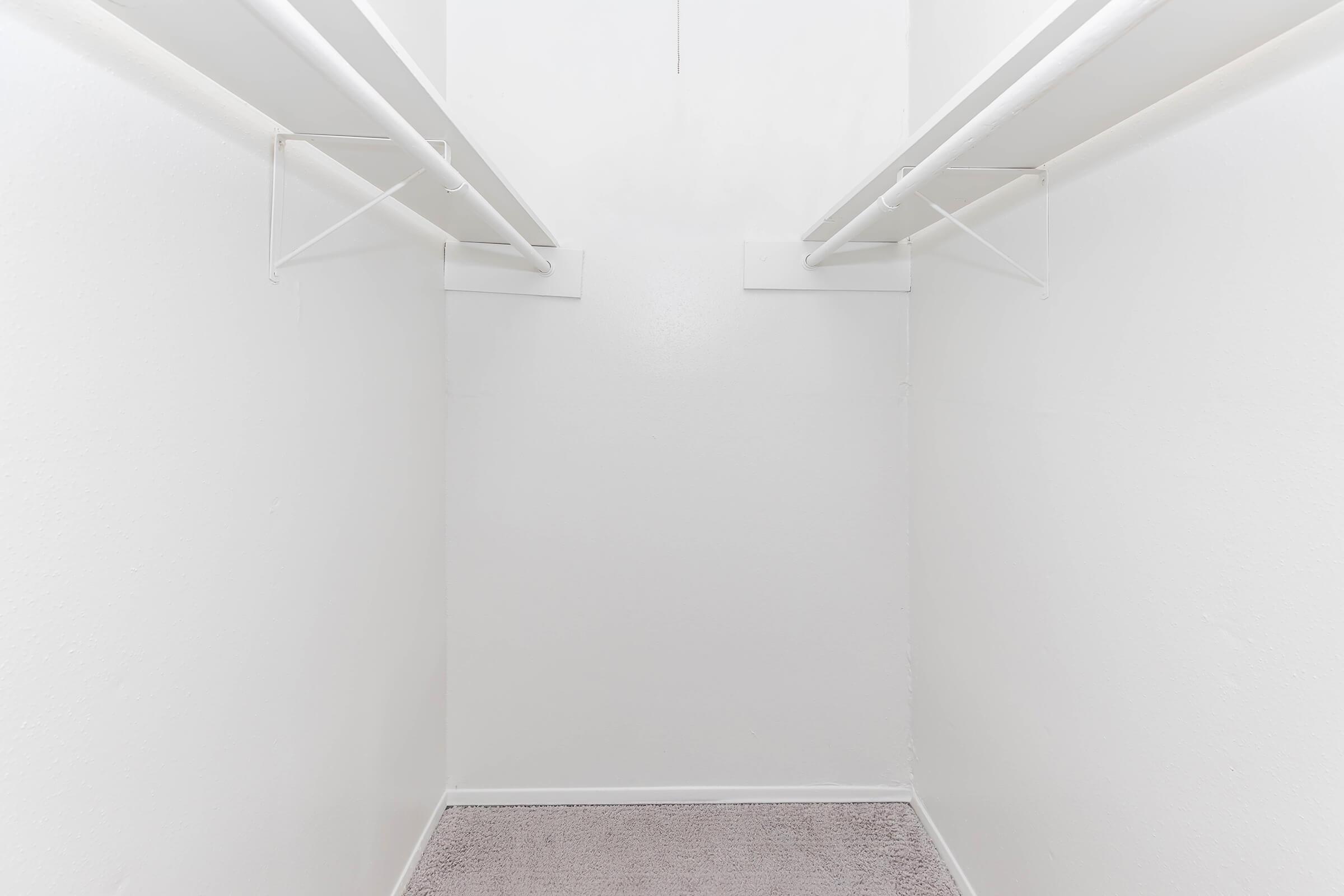 Empty white closet with two hanging rods on opposite walls, devoid of shelves or clothing. The floor is covered with a light-colored carpet, and the walls are plain and unadorned, creating a minimalist look.