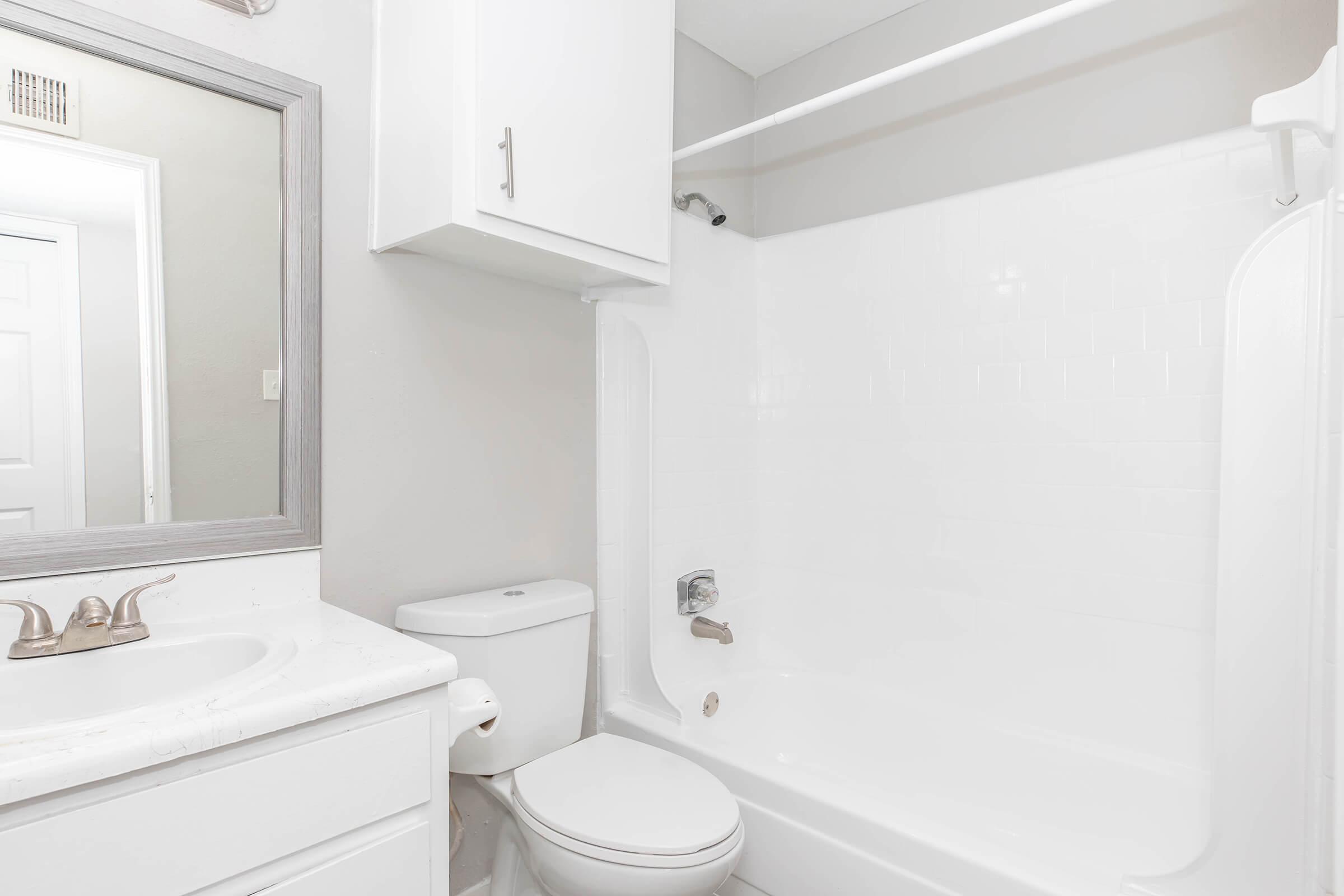 A clean and modern bathroom featuring a white bathtub with a shower, a white toilet, and a vanity with a sink. Light gray walls and a full-length mirror add to the spacious feel. There is also a cabinet above the sink for storage.