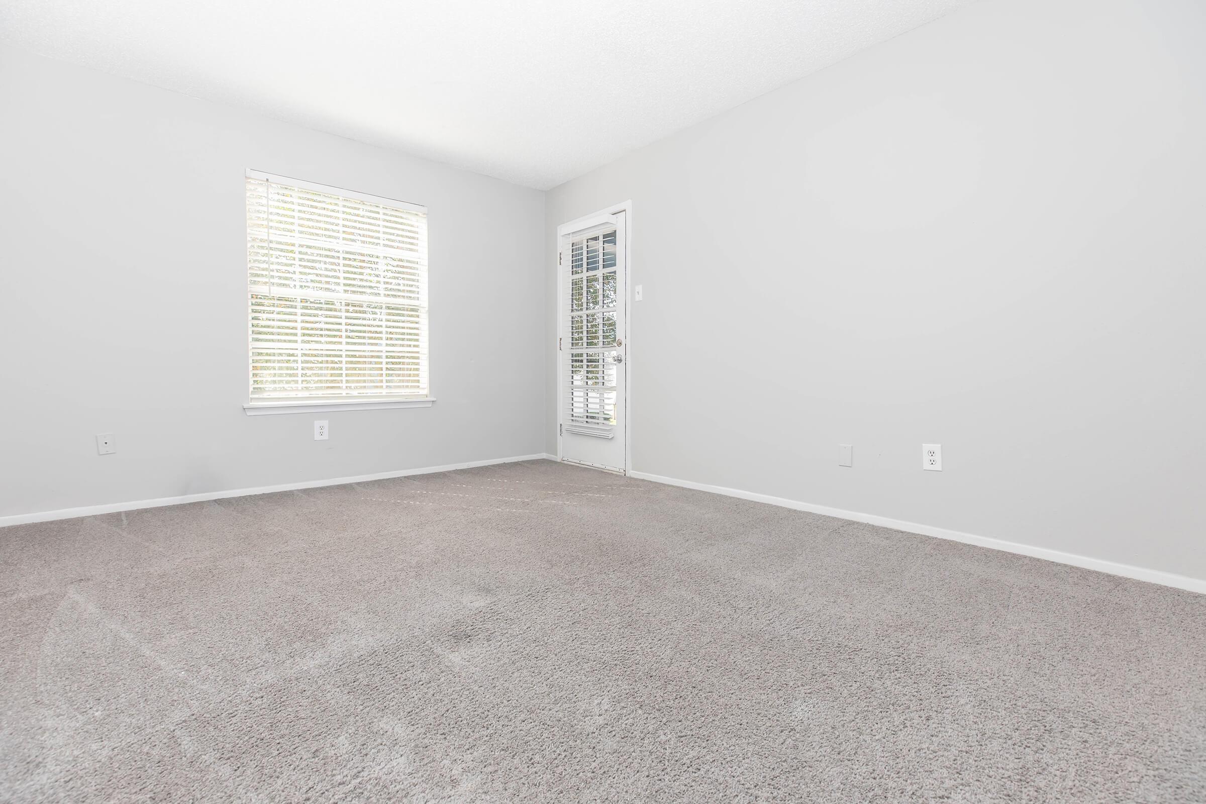 A spacious, empty room featuring light gray walls, a large window with white blinds allowing natural light, and a glass door leading outside. The floor is covered in beige carpet, creating a clean and neutral atmosphere, perfect for furnishing or personalizing.