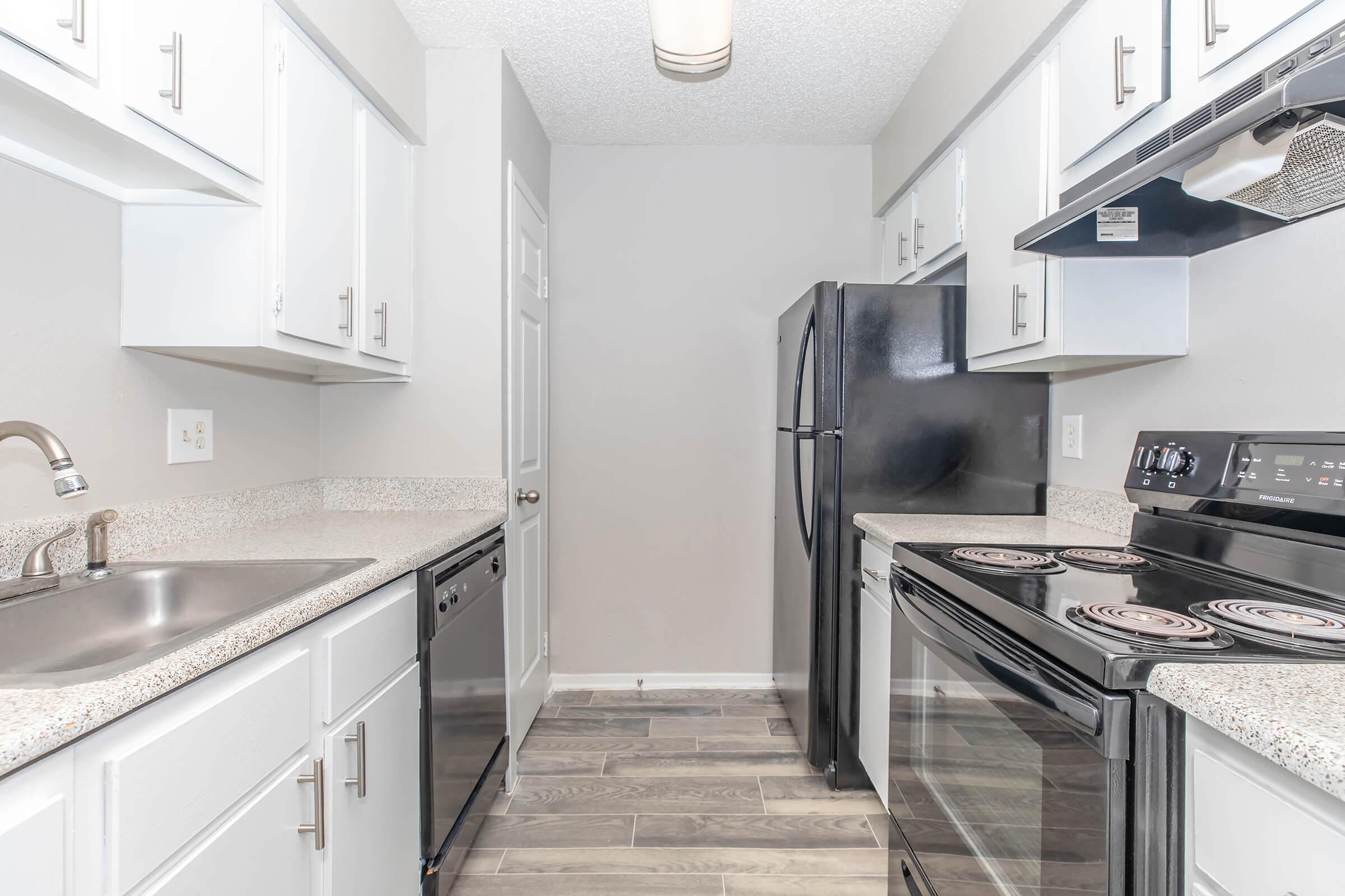 A spacious kitchen with modern appliances featuring white cabinets, a granite countertop, a double sink, and a black refrigerator. The flooring is a light wood look, and the kitchen includes an electric stove and oven with a black finish. Natural light illuminates the room from a ceiling fixture.
