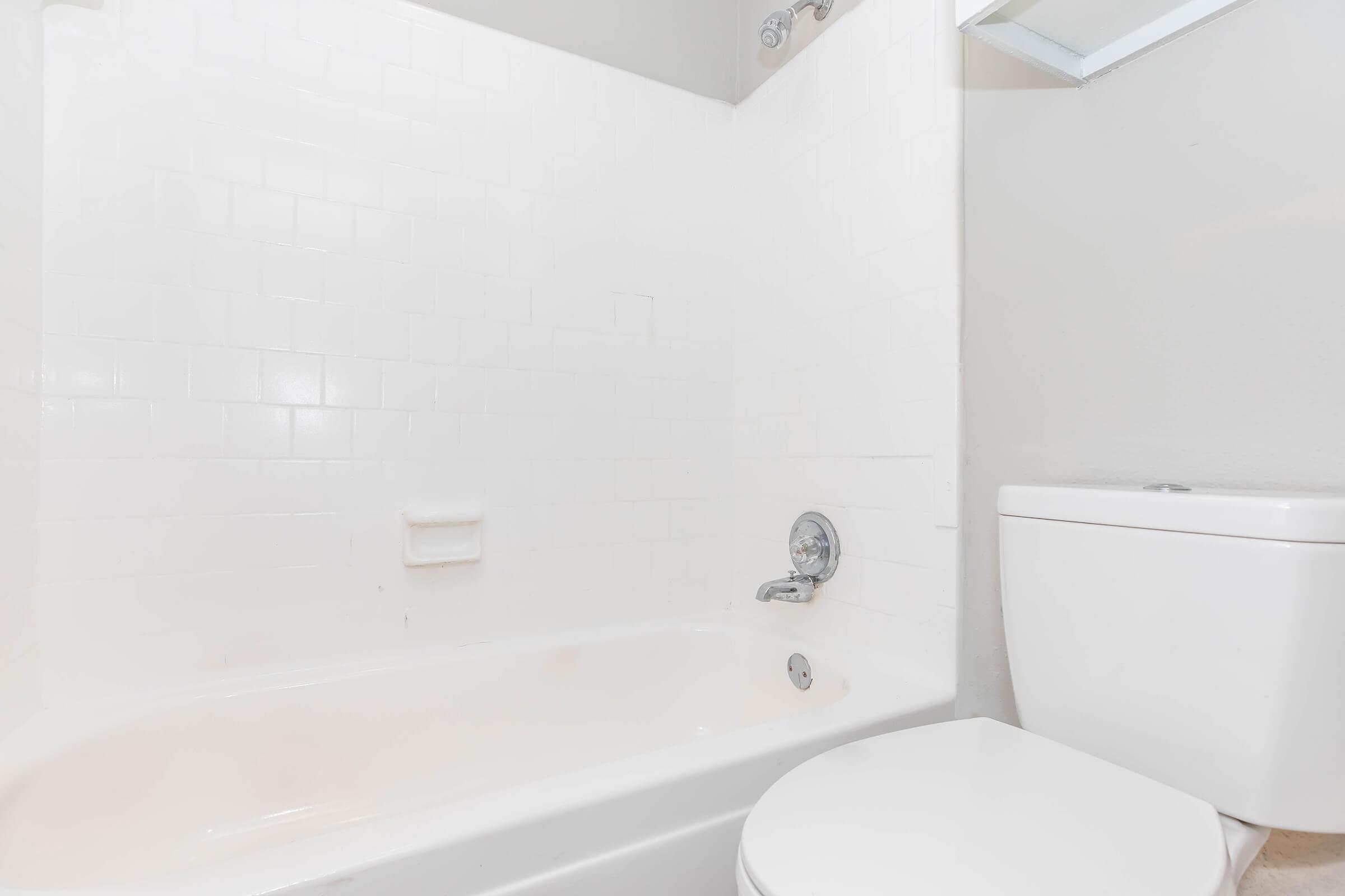 A clean, minimalistic bathroom featuring a white bathtub with a showerhead above, white tiled walls, and a modern toilet. The walls are painted light gray, enhancing the bright and spacious feel of the space. A small mirror is mounted on the wall above the sink area.