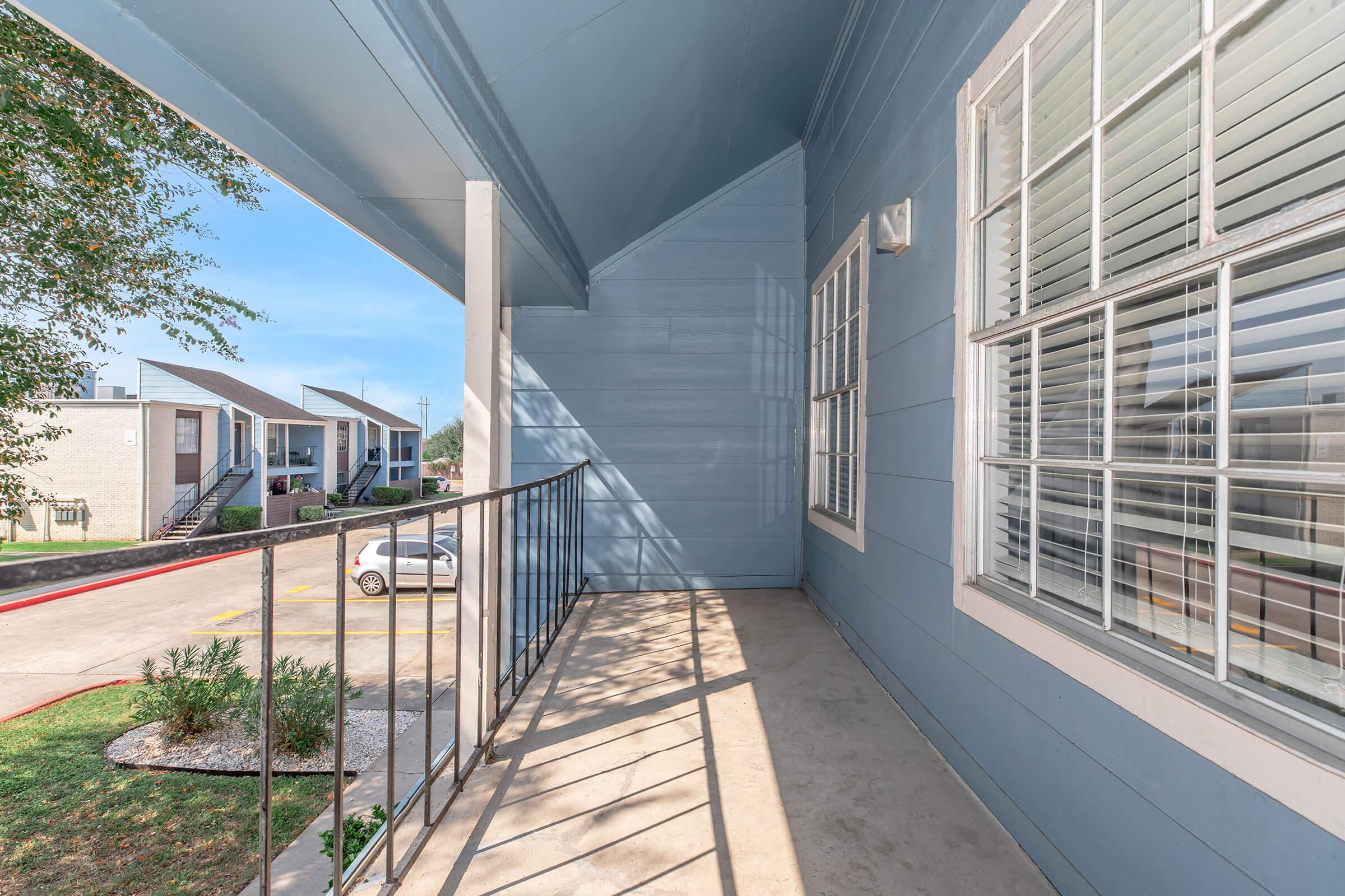 A spacious balcony with a concrete floor, framed by a metal railing, featuring light blue walls. The view shows a grassy area, parked cars, and apartment buildings in the background under a clear blue sky. Sunlight casts gentle shadows, creating a serene outdoor space.