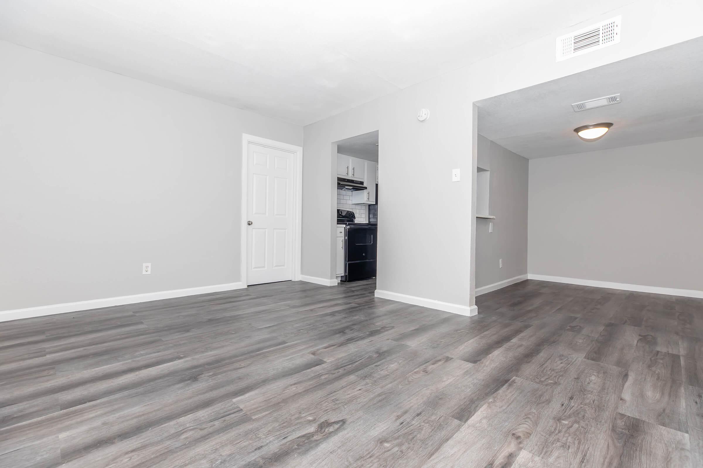 A spacious, empty living area with light gray walls and wood-style flooring. To the left, there's a closed white door, and a doorway leads into a kitchen area equipped with modern appliances. The room is well-lit, creating a clean and inviting atmosphere.