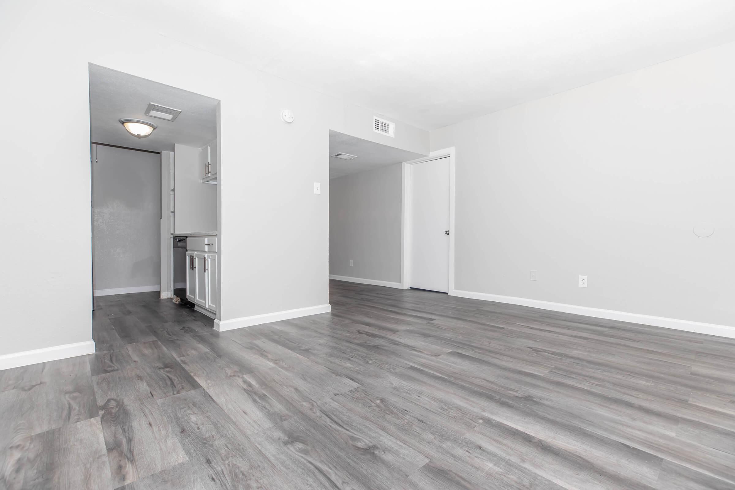 A spacious, empty room with light gray walls and modern flooring. There is an opening leading to a small kitchen area on the left, and a closed door on the right. The room is brightly lit, creating a clean and airy atmosphere.