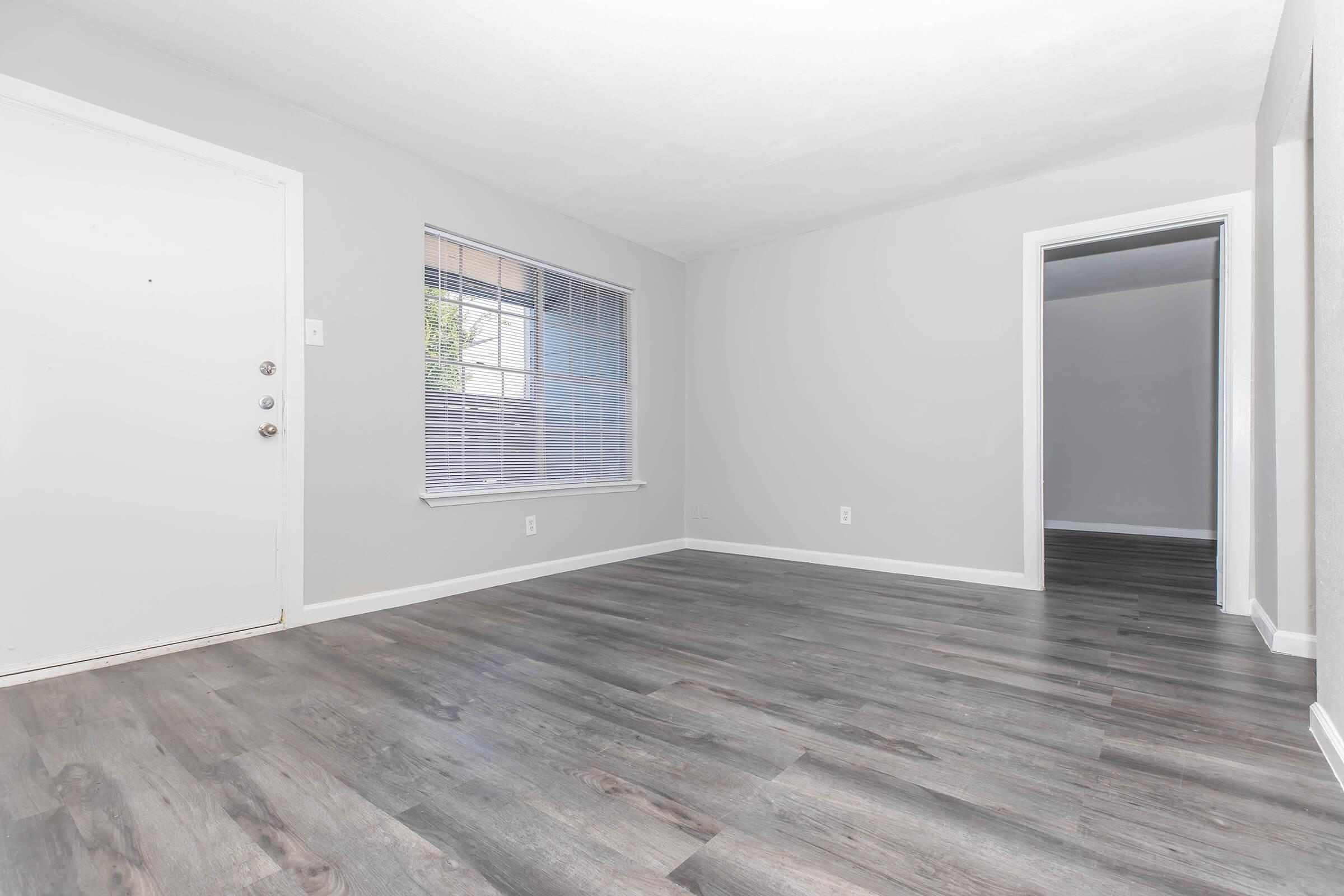 Light-filled room with gray walls and laminate flooring. A white door leads to the outside on the left, and a window with blinds is located on the front wall. An entryway leads to another room on the right side, indicating an open and spacious layout.