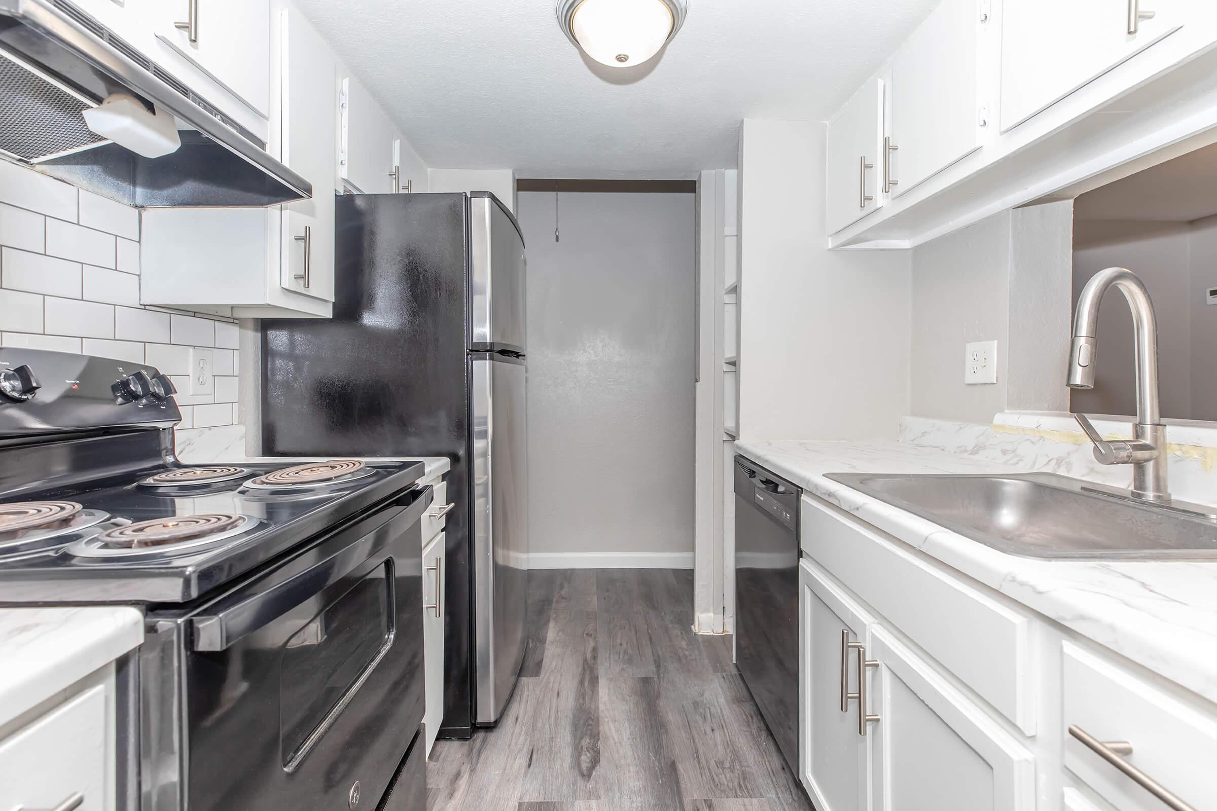 A modern kitchen featuring white cabinetry, a black stove and refrigerator, stainless steel sink, and marble countertop. The floor is a light wood laminate, and there's a doorway leading to another space, adding a bright and airy feel to the room.