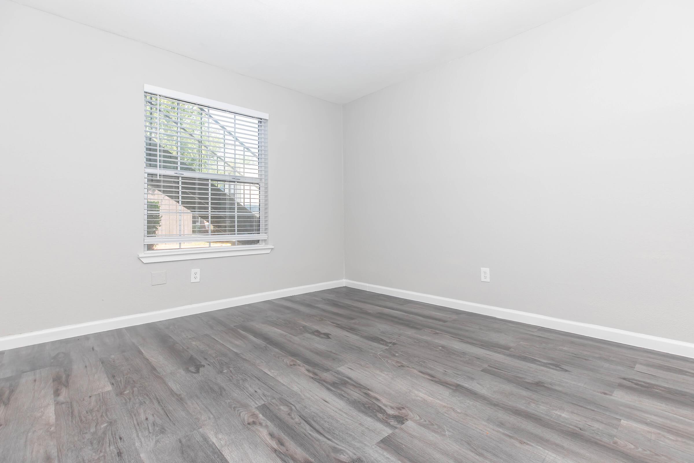 A well-lit empty room with light gray walls, a window featuring blinds, and a gray wooden floor. The room appears spacious, showcasing the simplicity of the design and ample natural light coming through the window.