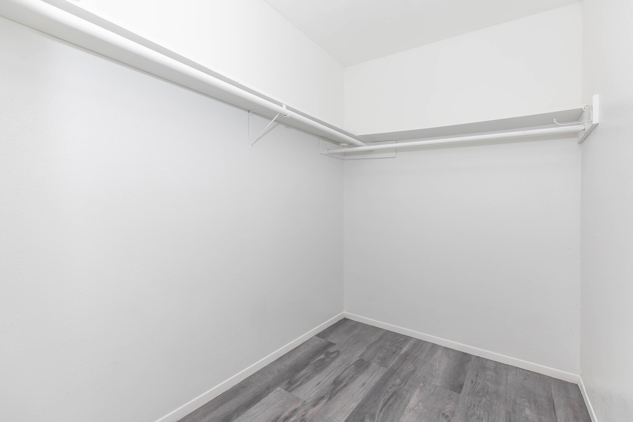 Empty closet with white walls and a wooden floor. There is a single hanging rod along the top for clothes, with no shelves or additional storage present. The overall atmosphere is clean and minimalistic, creating a spacious feel.