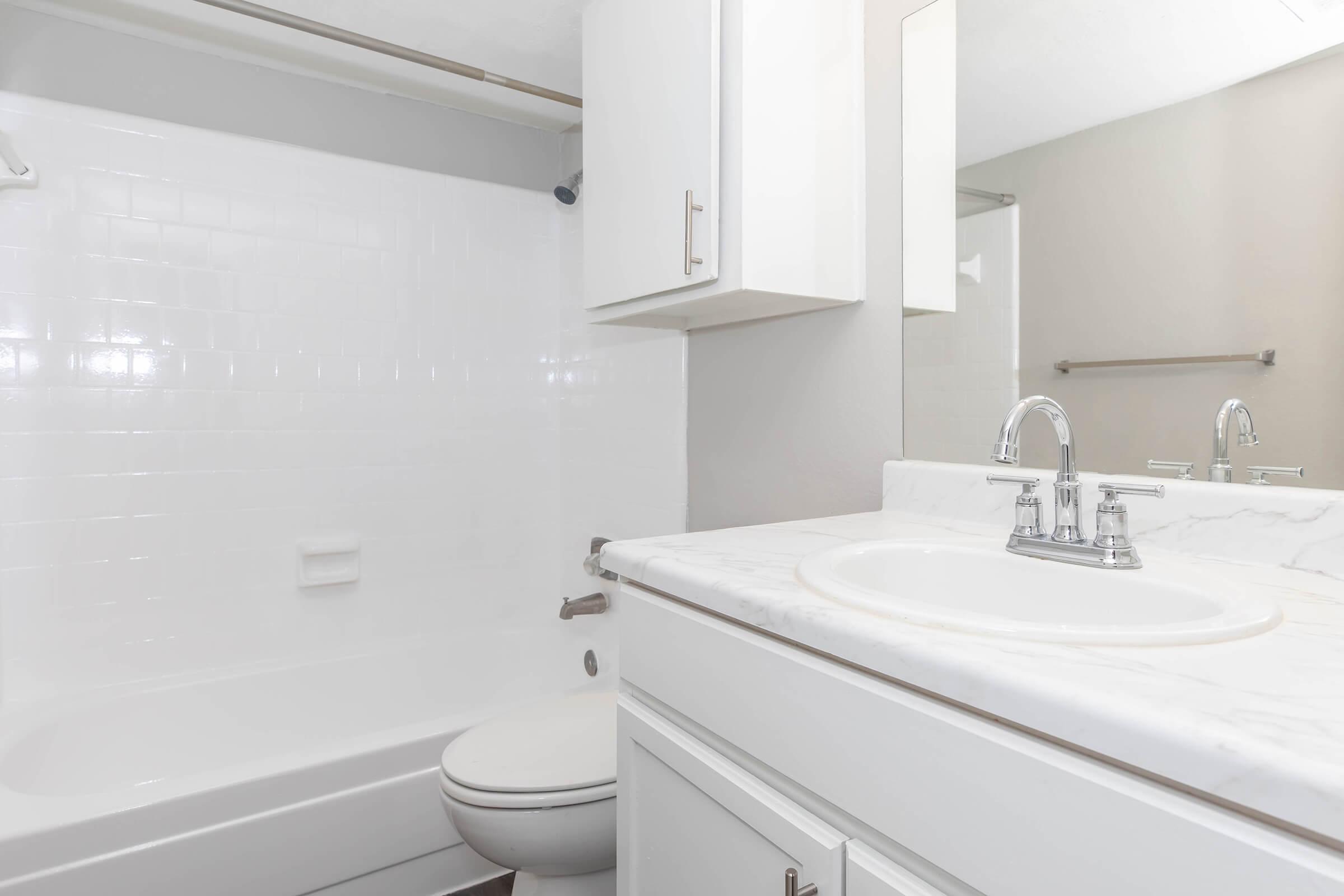 A clean, modern bathroom featuring a white bathtub with a shower, a white vanity with a marble countertop, and a mirror. The walls are painted in a light color, and there is a towel bar and a cabinet for storage. The overall design is simple and functional.