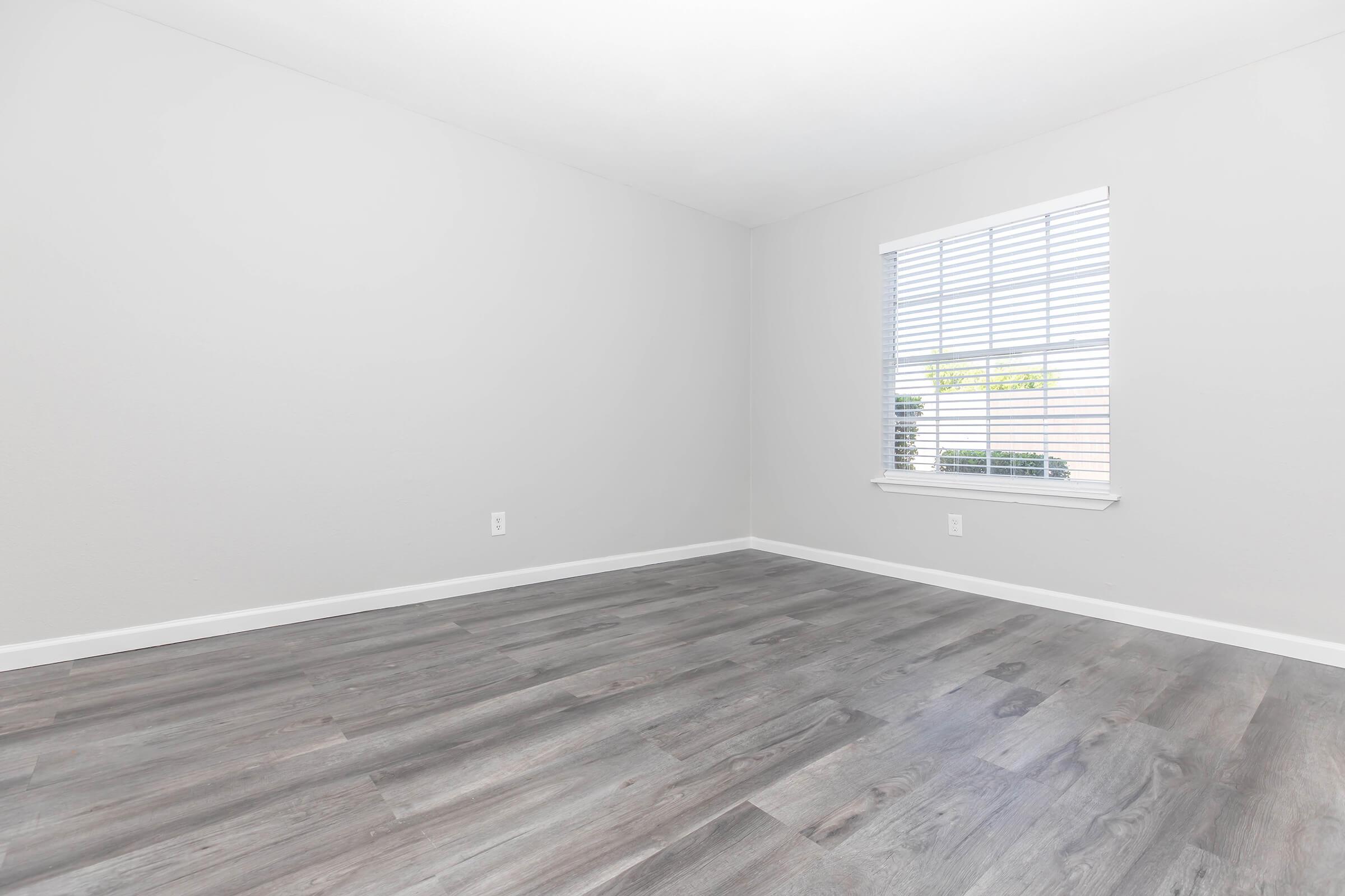 Empty room with gray walls and light-colored wood flooring. A window with white blinds allows natural light to enter. No furniture or decorations present, creating a minimalist and spacious atmosphere.