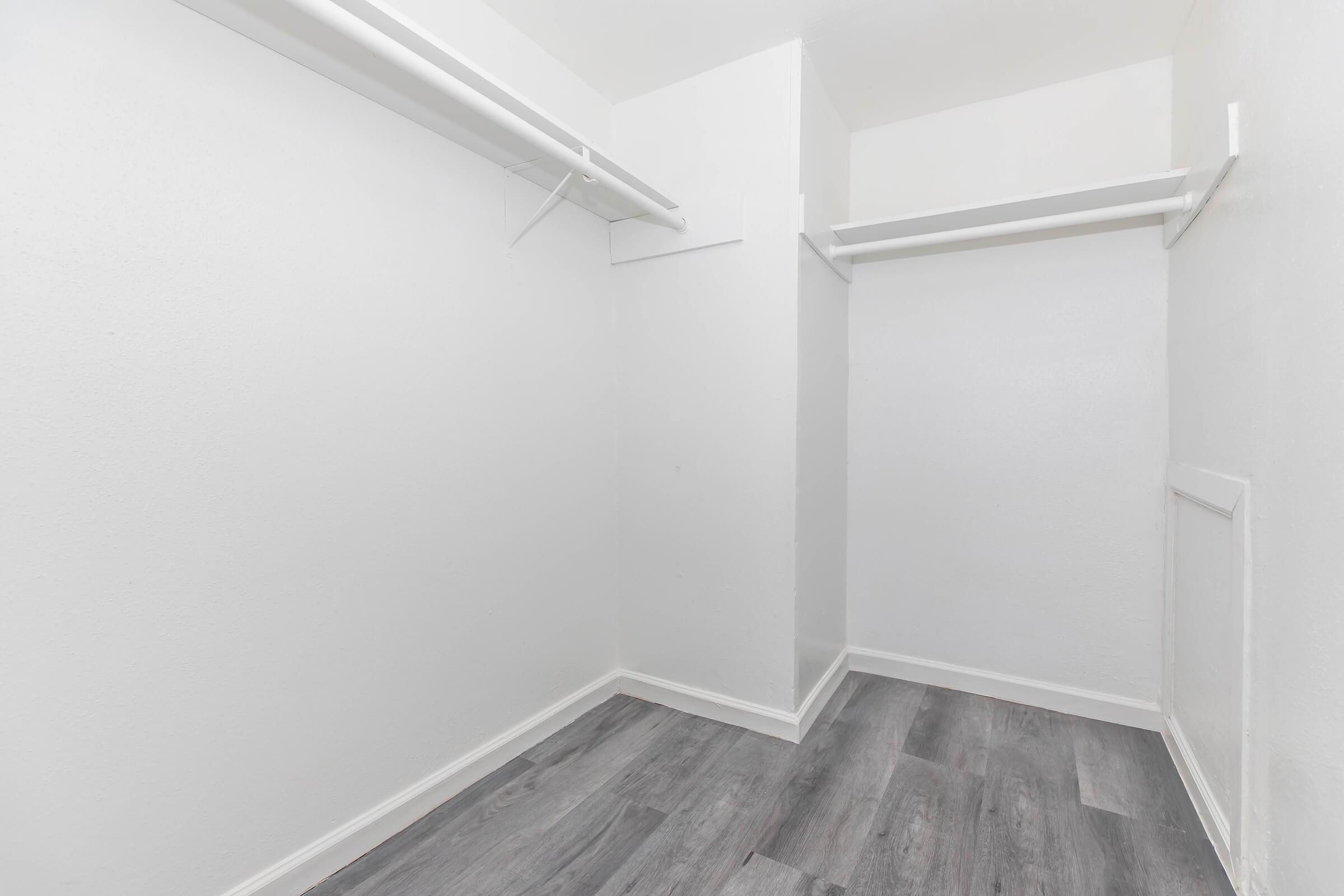 Empty closet with white walls and a light wood-like floor. It features two hanging rods on the walls, providing storage space, and has no shelves or items inside. The overall space appears clean and minimalistic, suitable for organizing clothes or accessories.