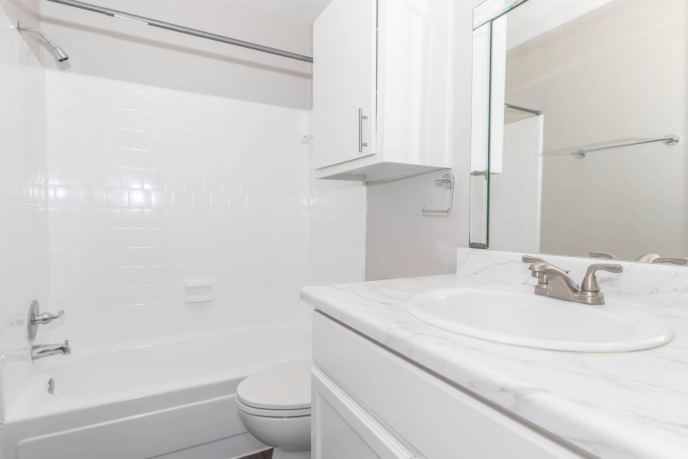A clean and modern bathroom featuring a white tiled bathtub, a sink with a marble countertop, and a mirror above the sink. The walls are painted in a light color, and there are white cabinets for storage. The overall design is simple and well-maintained, providing a bright and inviting atmosphere.