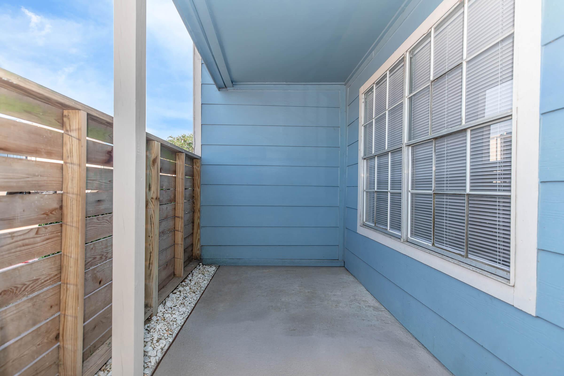 A small, empty patio area with a concrete floor, blue wooden walls, and a fenced side. The sunlight casts soft shadows, and the space is partially shaded by a roof overhang. A window with multiple panes offers a view outside.