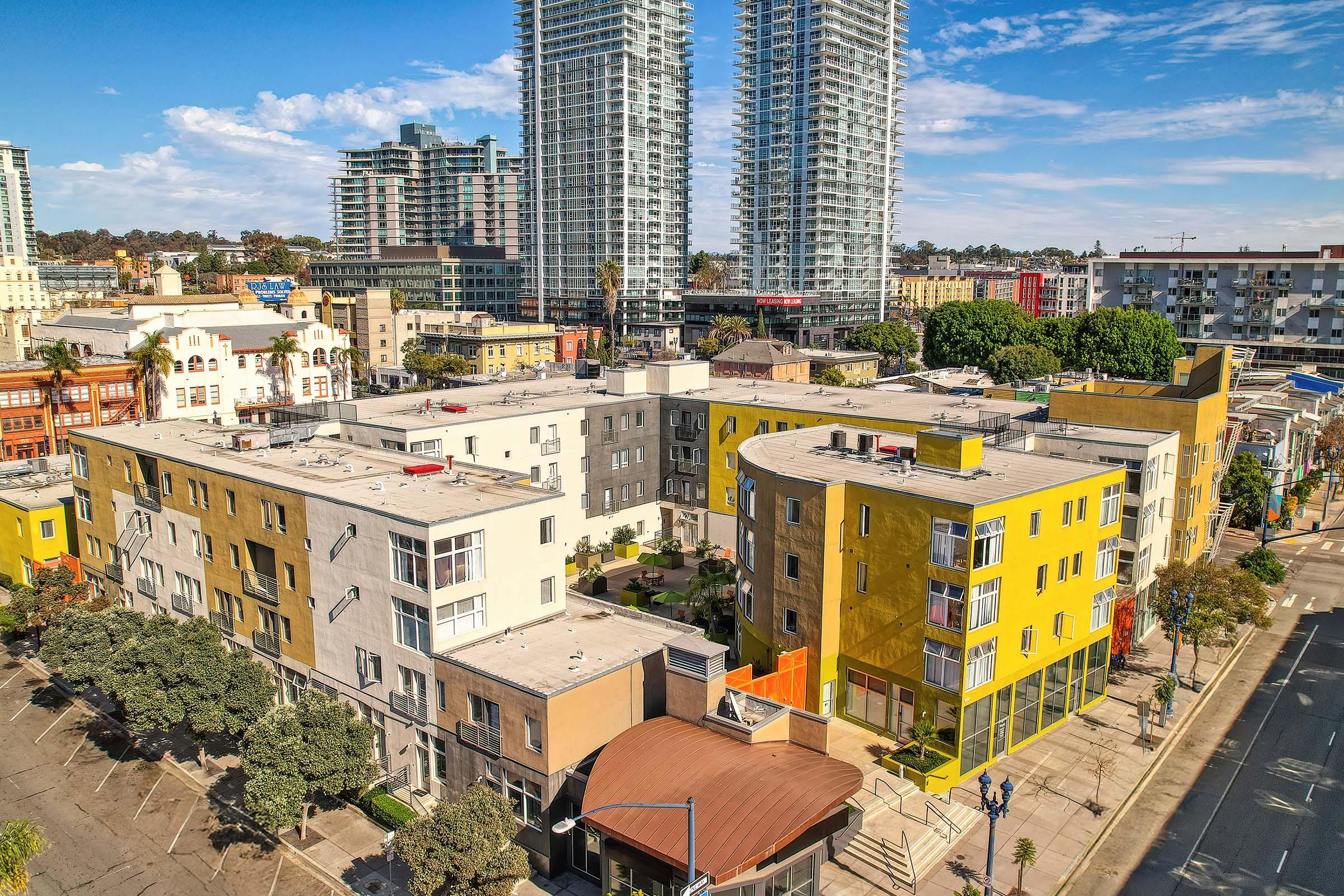 Aerial view of an urban area featuring modern high-rise buildings alongside colorful mid-rise apartments. The scene includes well-maintained landscaping, street access, and a clear blue sky, reflecting a vibrant city environment.