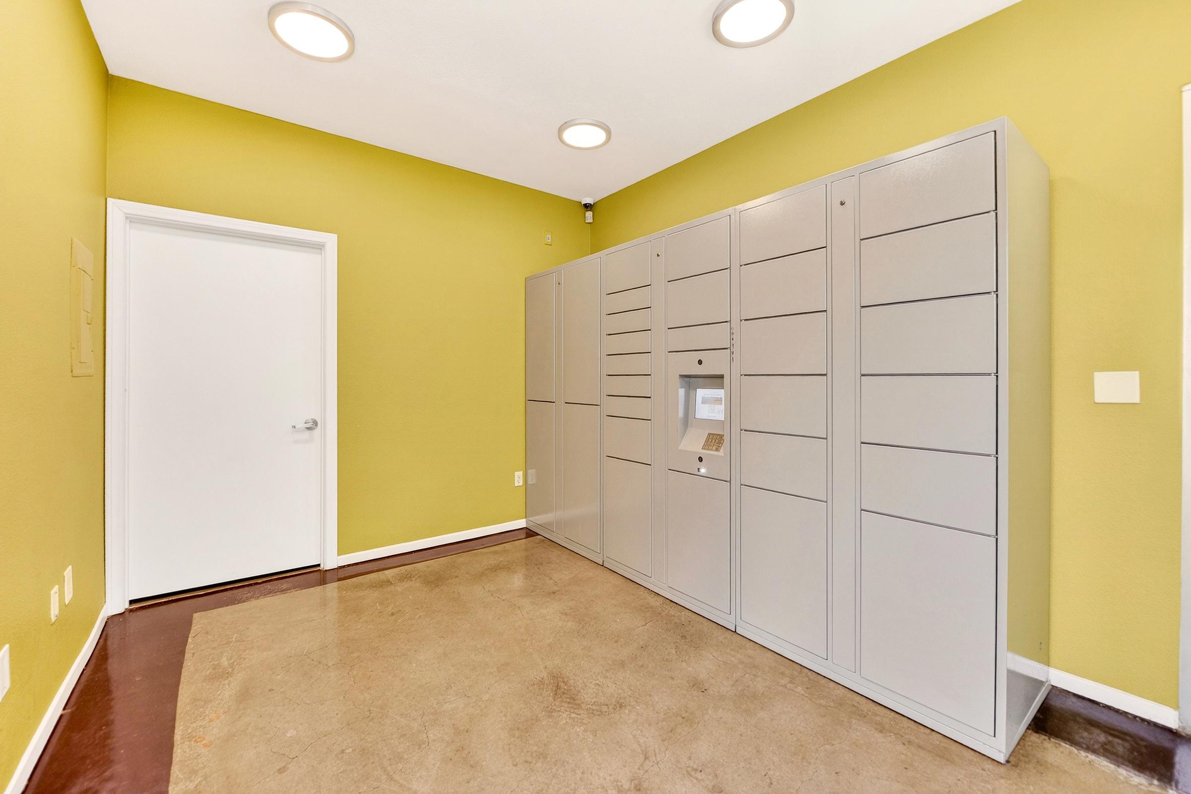 A modern, empty room with light green walls and a concrete floor. On one side, there is a row of gray lockers with a central touchscreen interface, indicating a package delivery or storage area. A white door is visible on the opposite wall. The space is well-lit with circular ceiling lights.