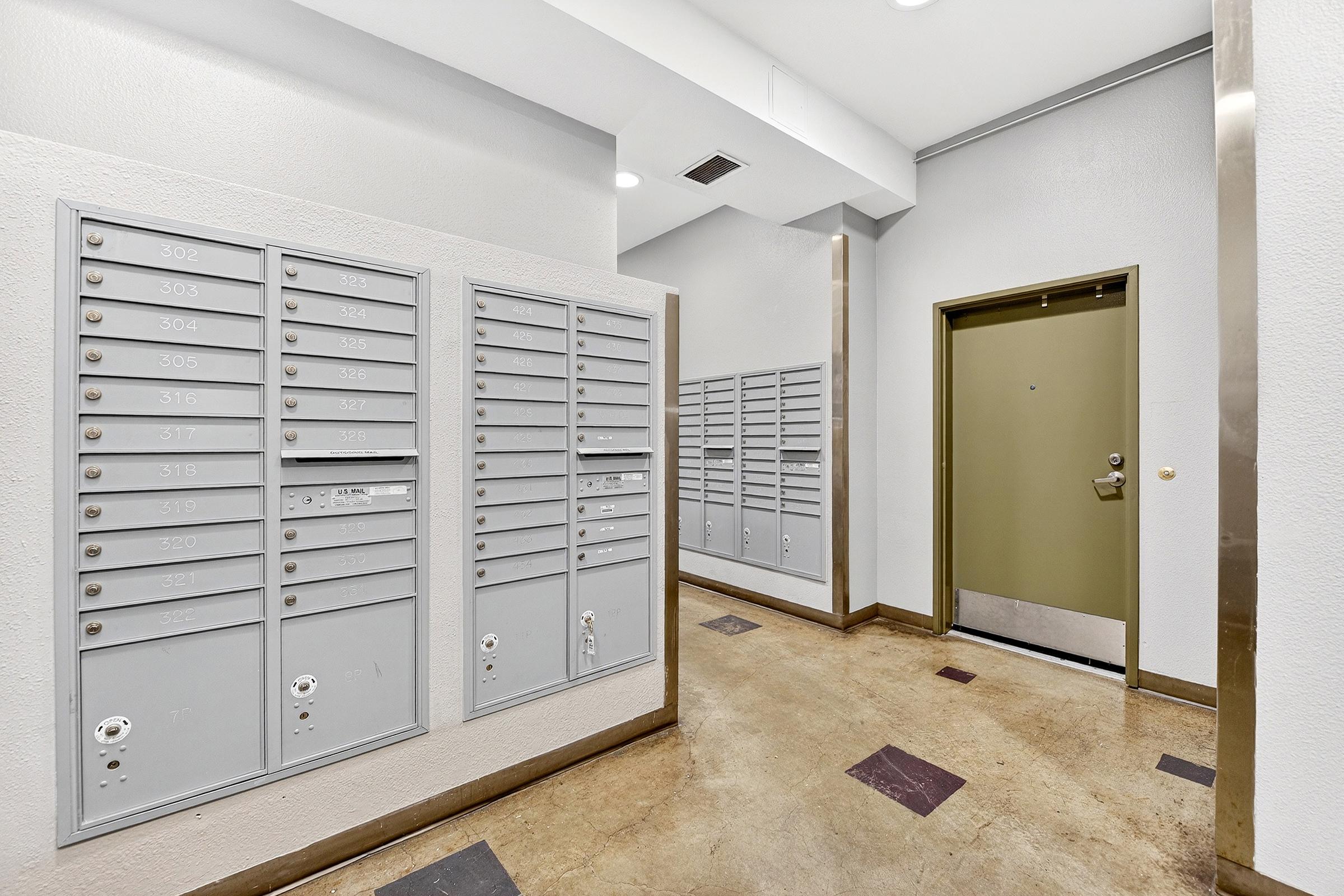 A hallway with multiple gray postal mailboxes mounted on the wall, alongside a green door leading to an apartment. The floor is made of polished concrete with square tiles. The walls are painted light gray, and there are overhead lights illuminating the space.