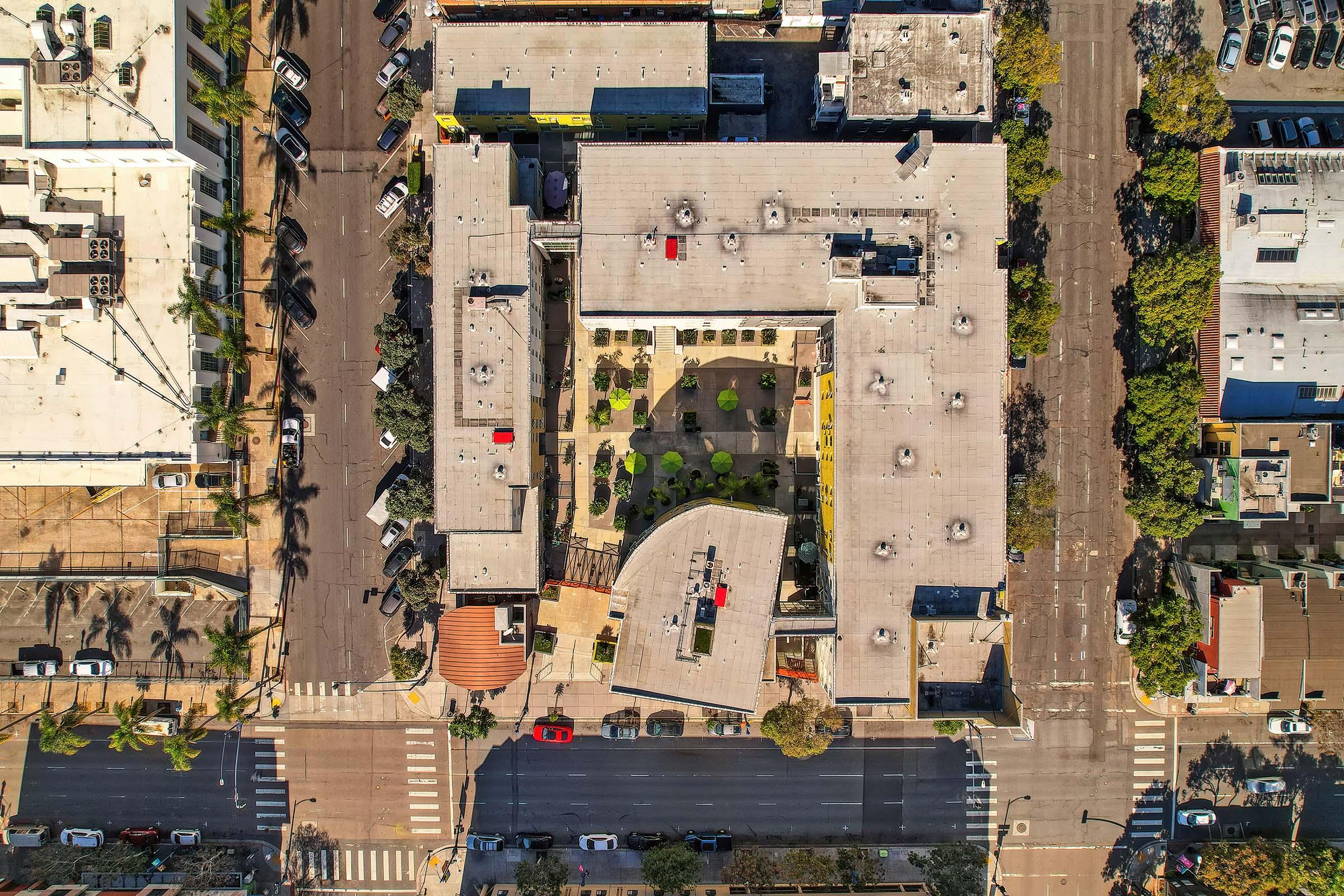 Aerial view of a city block featuring a modern building complex with a central courtyard. The area is surrounded by streets filled with parked cars and trees, showcasing urban landscaping. The layout includes various rooftops and an open plaza with greenery, highlighting a blend of architecture and nature in an urban setting.