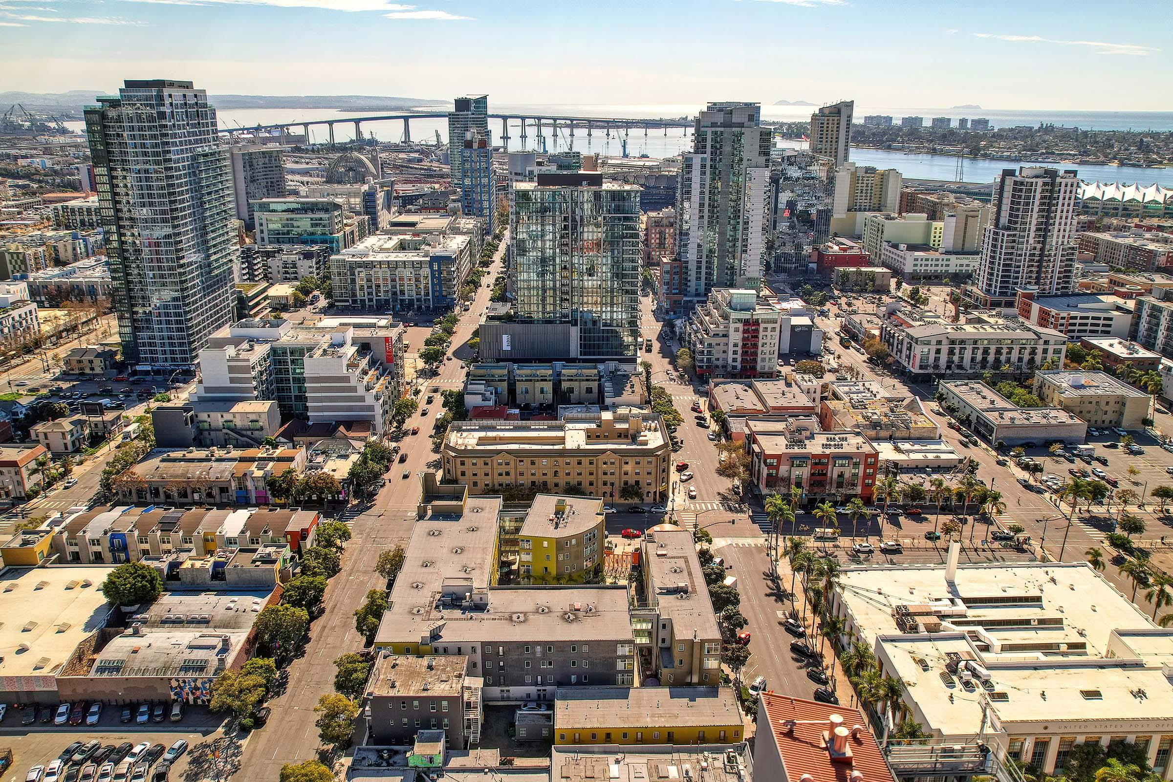 Aerial view of a bustling urban area featuring a mix of modern high-rise buildings and lower structures, with streets lined by trees and cars. The skyline includes a bridge in the background and is set against a clear blue sky. This urban landscape showcases a vibrant city atmosphere.