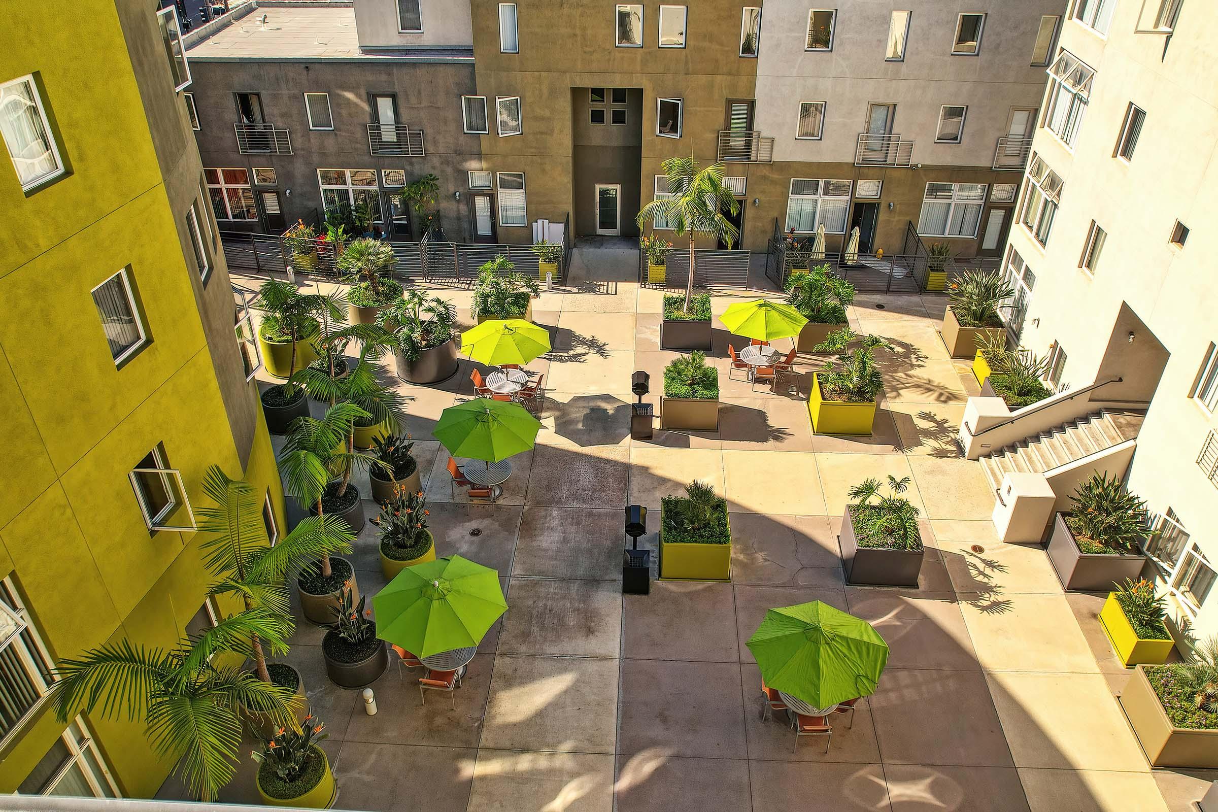 A courtyard view of a residential complex featuring several green umbrellas over outdoor seating areas. The space is surrounded by modern apartment buildings, potted plants, and a few palm trees, creating a vibrant and inviting atmosphere. Sunlight casts shadows on the pavement.