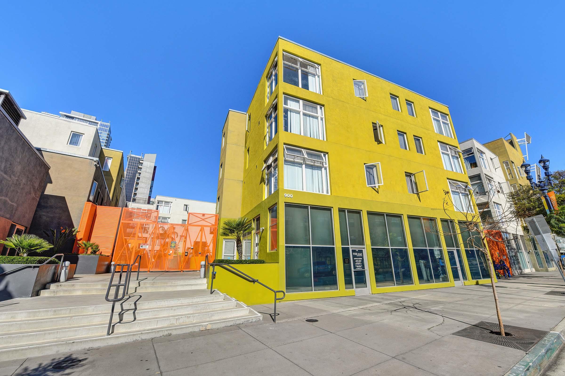 A modern, colorful building with a bright yellow exterior and large windows, featuring an orange entrance. The structure has a contemporary design, surrounded by a paved walkway and a few palm trees. Clear blue skies above enhance the vibrant colors of the building.